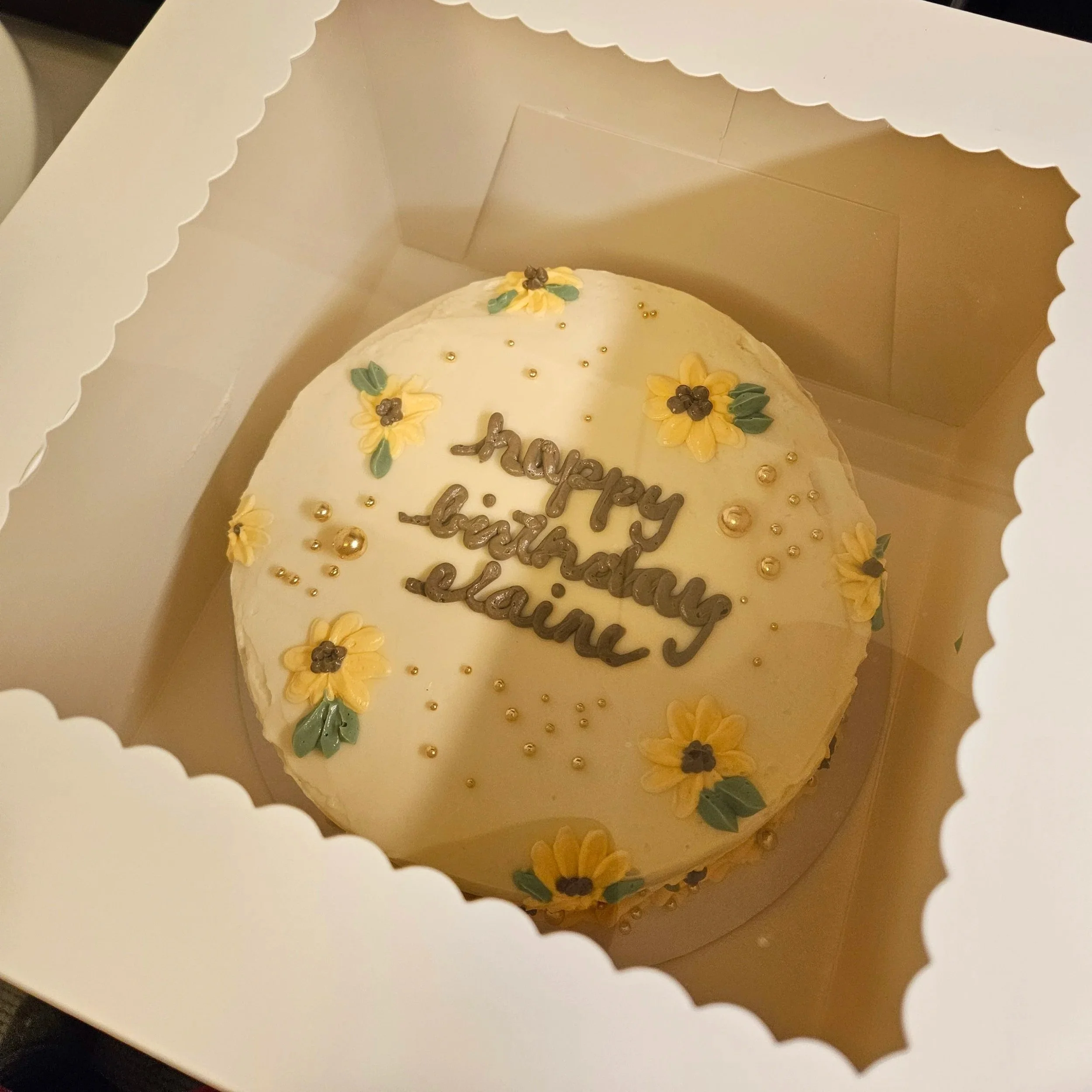 Close-up of a white cake inside a box with scalloped edges, decorated with yellow flowers, green leaves, gold beads, and a silver topper that spells out 'happy birthday'.