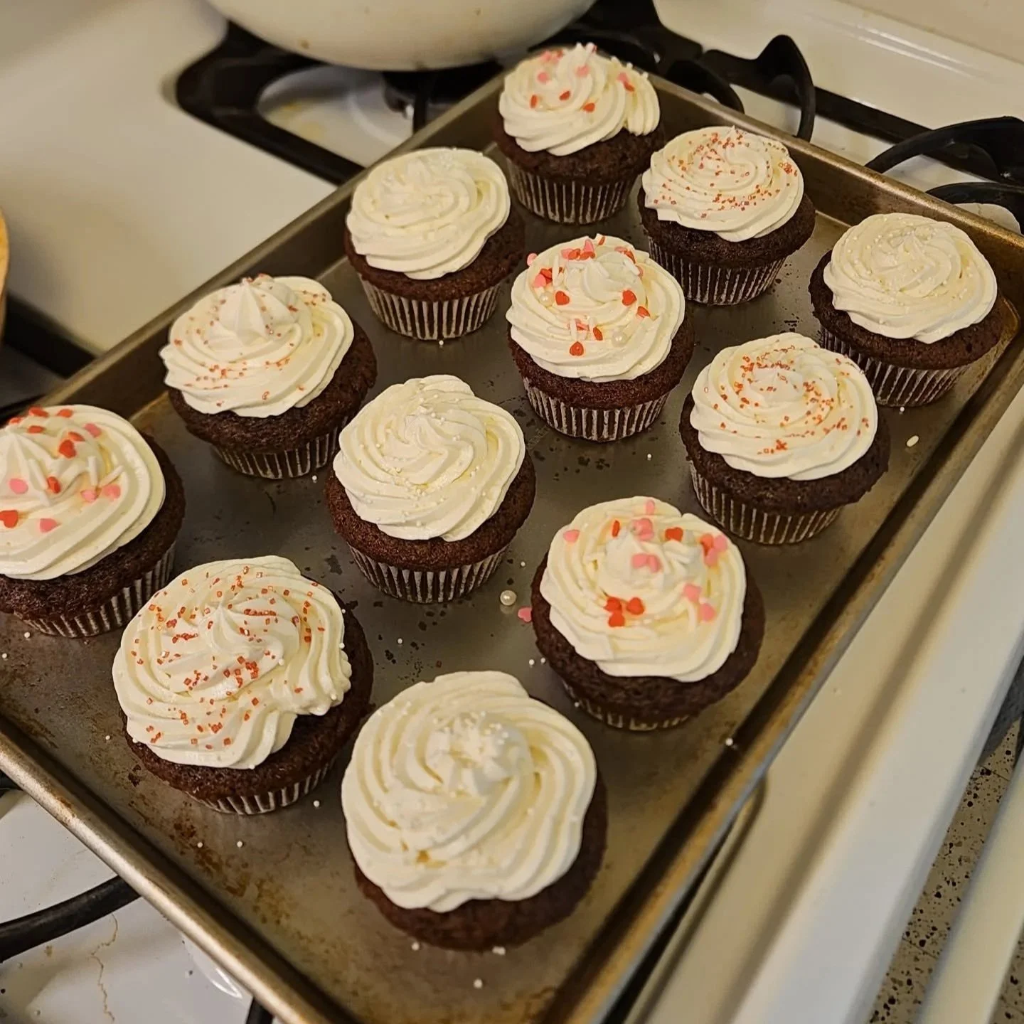 Chocolate cupcakes with white frosting and colorful sprinkles on a baking sheet in a kitchen.