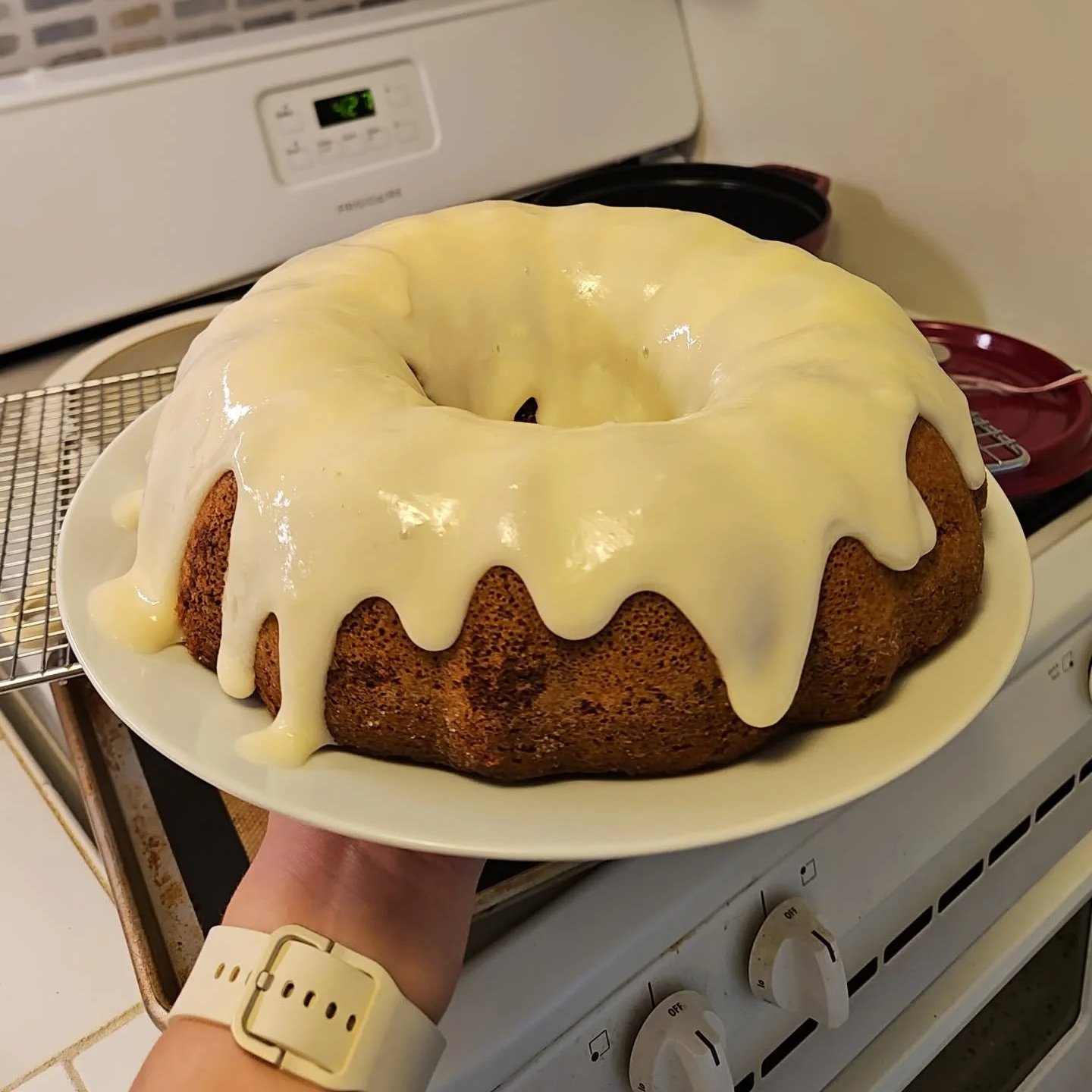 A bundt cake with yellow icing dripping over the sides being held on a white plate in a kitchen.