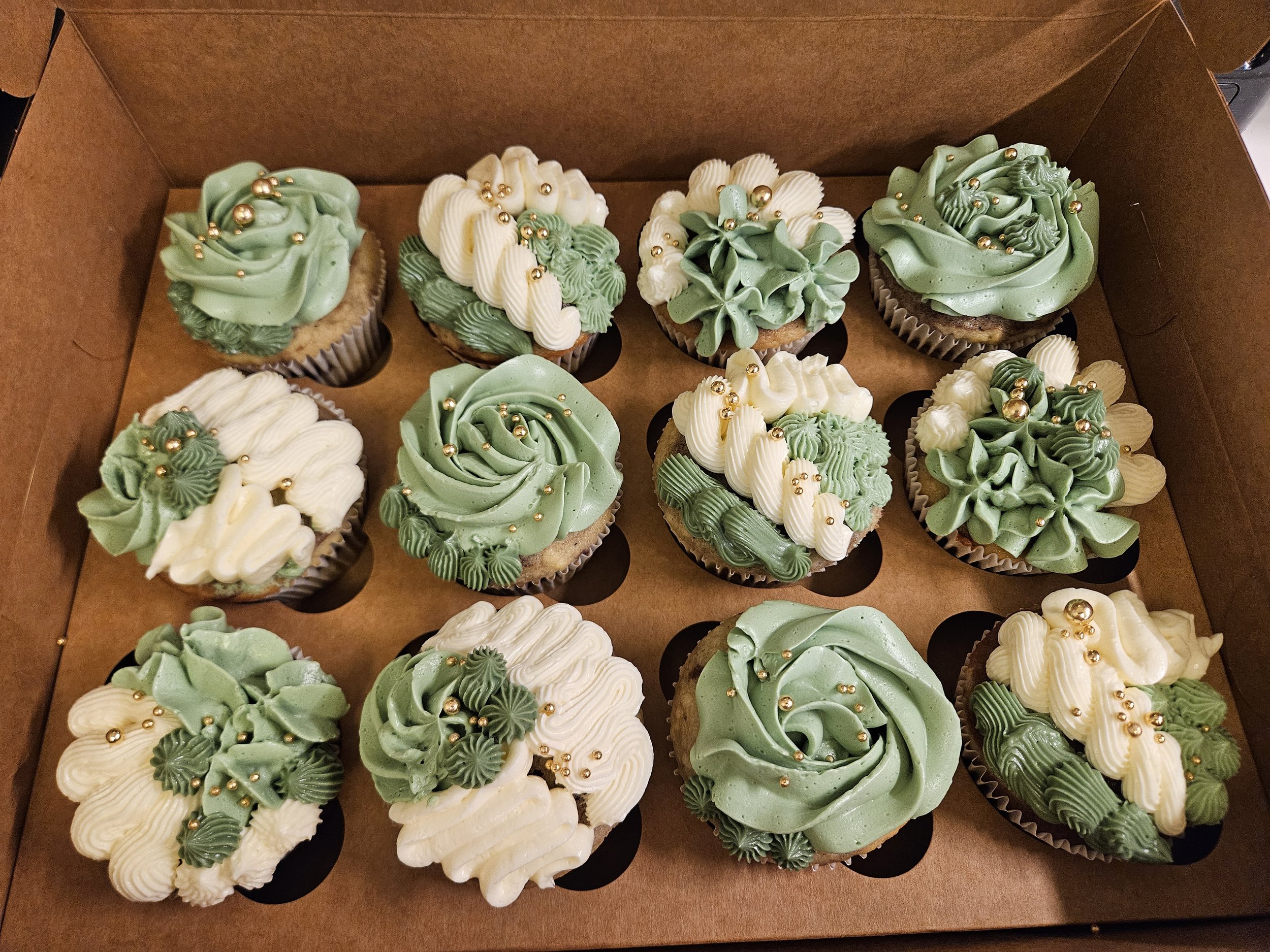 A box of decorated cupcakes with green and white buttercream frosting swirls topped with gold edible beads.