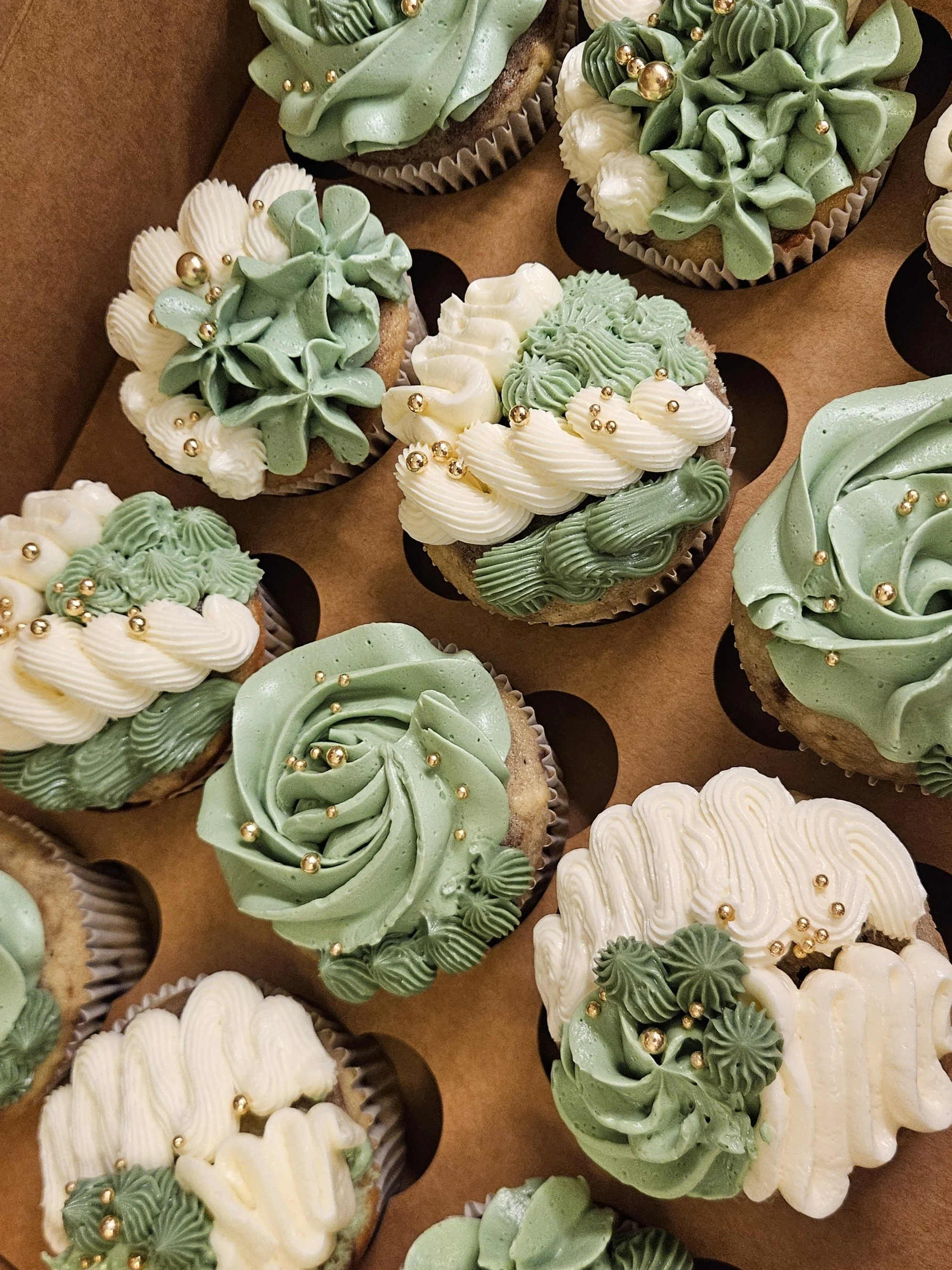 Assorted cupcakes decorated with green and white frosting, gold sugar pearls, arranged on a brown paper-lined tray.