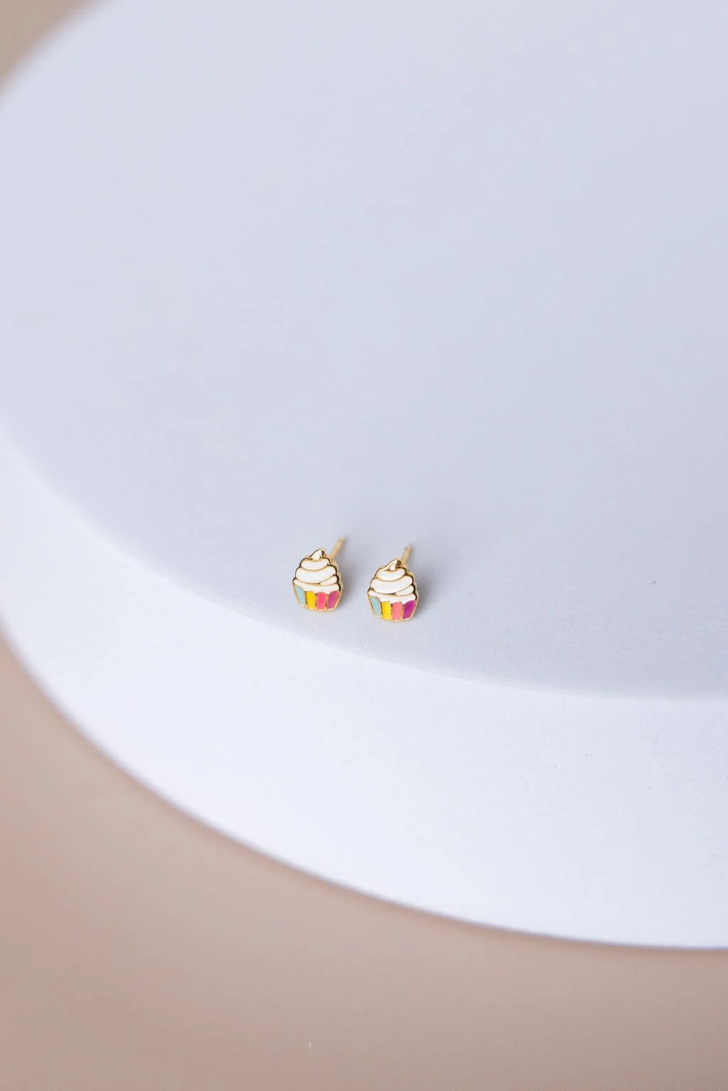 A pair of cupcake-shaped earrings with colorful frosting and rainbow-colored base on a white circular surface.