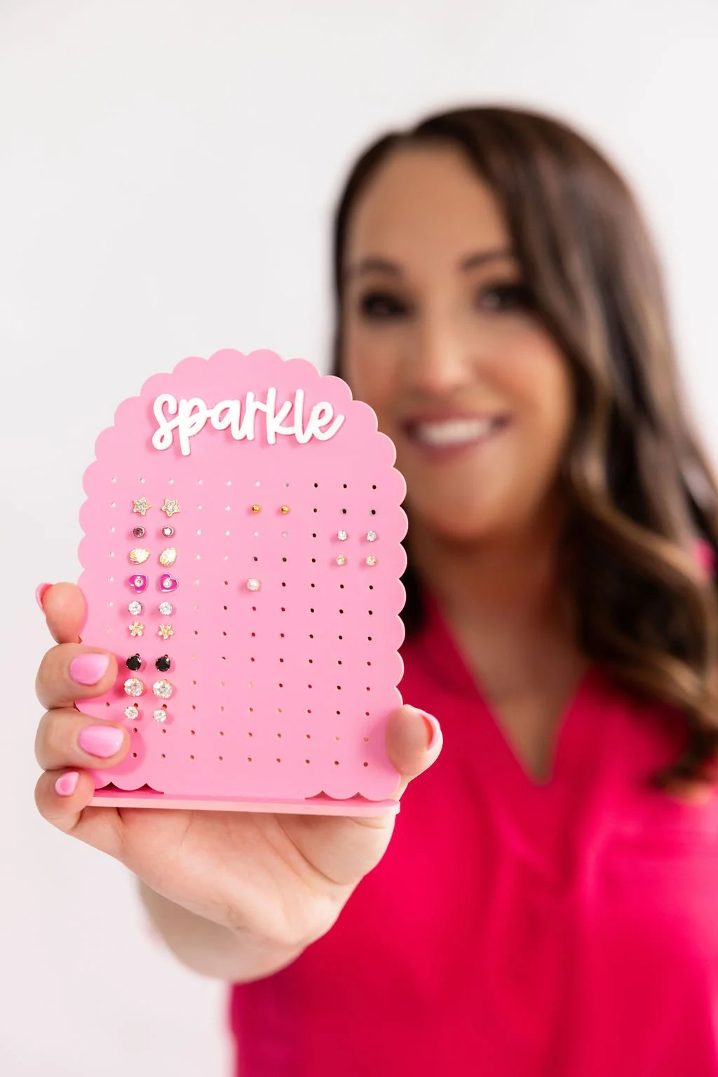 A woman holding a pink display stand with various small earrings, smiling in the background.