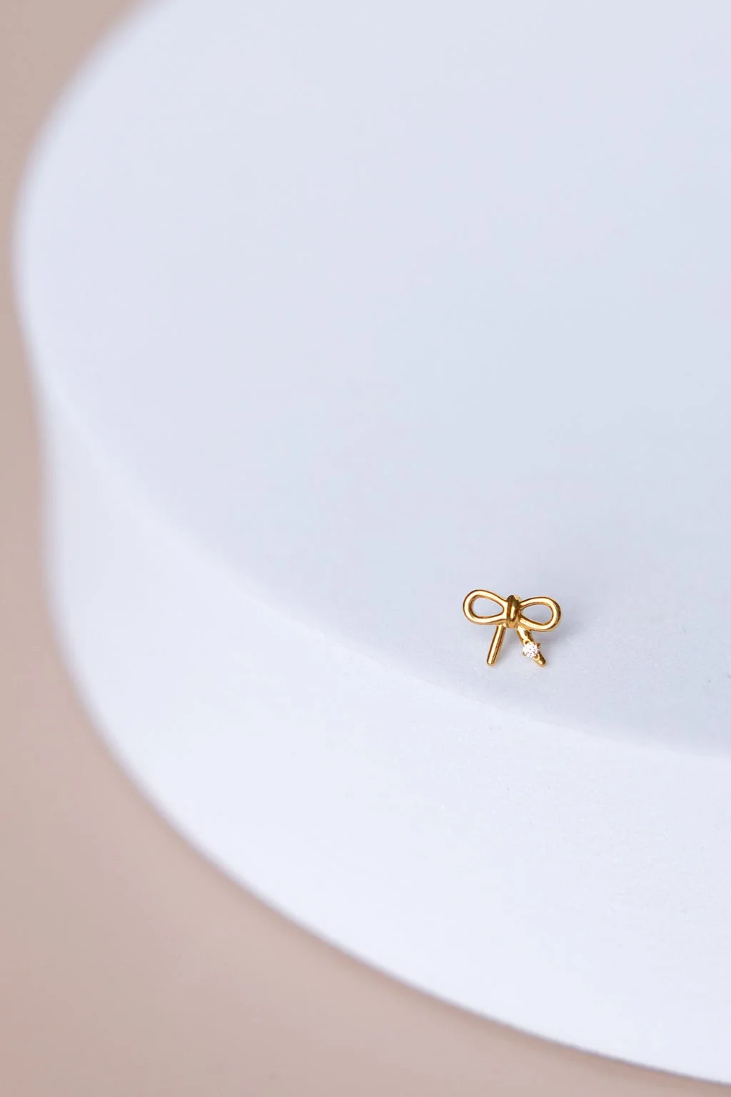 A small gold bow-shaped earring with a tiny gemstone, resting on a white circular surface.