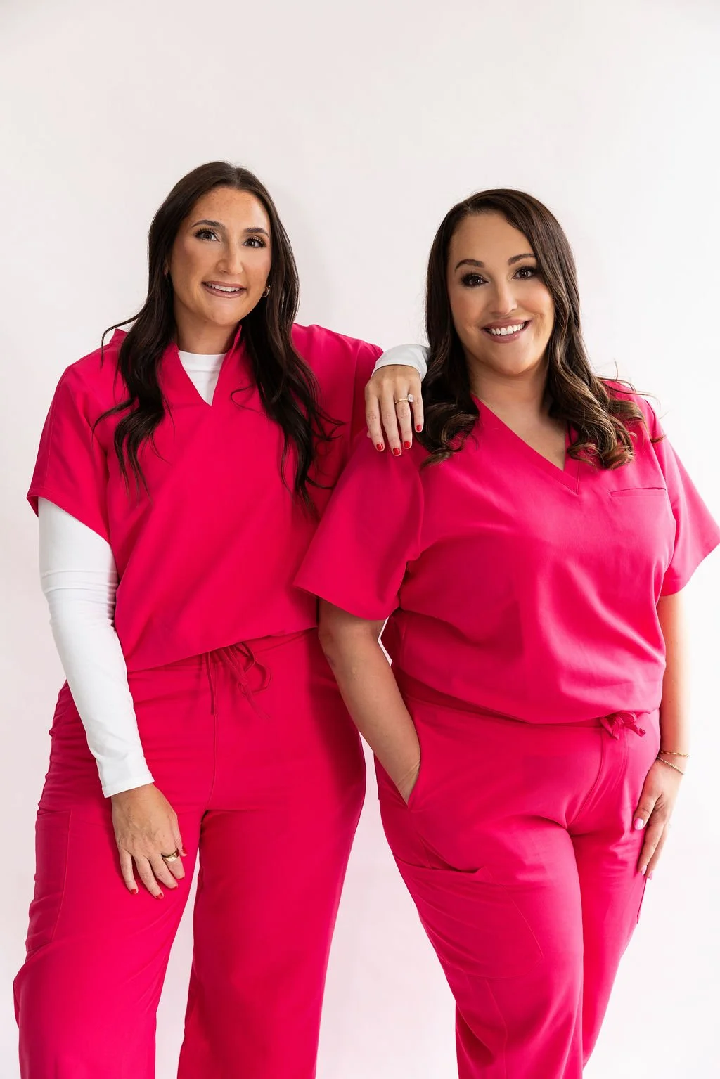 Two women dressed in pink medical scrubs posing against a white background, smiling at the camera. One woman has her arm resting on the other's shoulder.