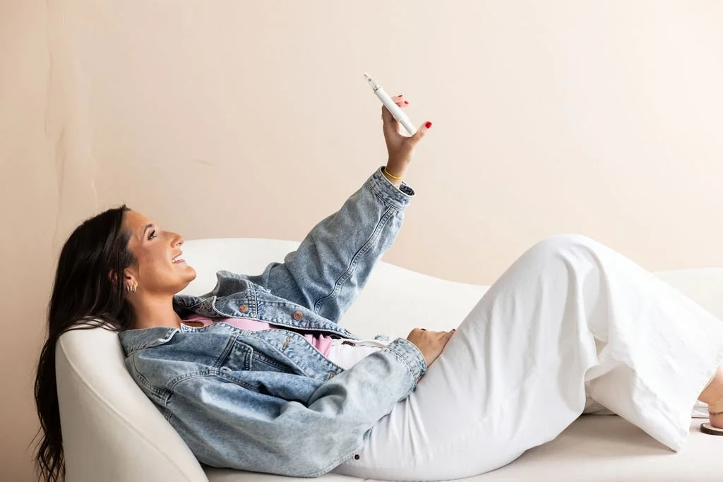 Woman lying on a white couch, holding an ear piercing device, with her phone, wearing a denim jacket and white pants.