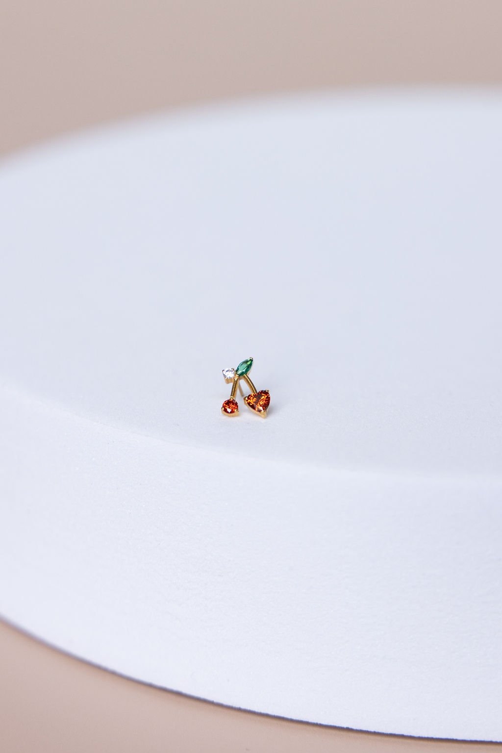 A small, colorful earring with a design resembling cherries with green, orange, and clear stones, placed on a white surface.