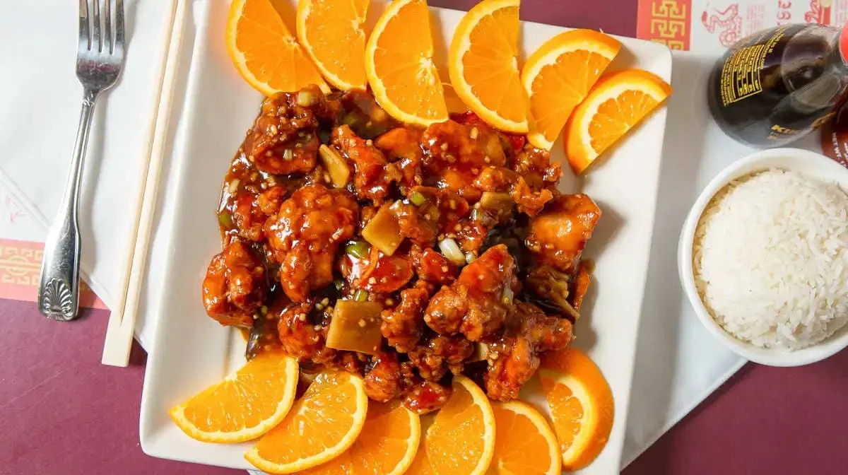 Chinese orange chicken with orange slices, white rice, and a soy sauce bottle on a white tray.