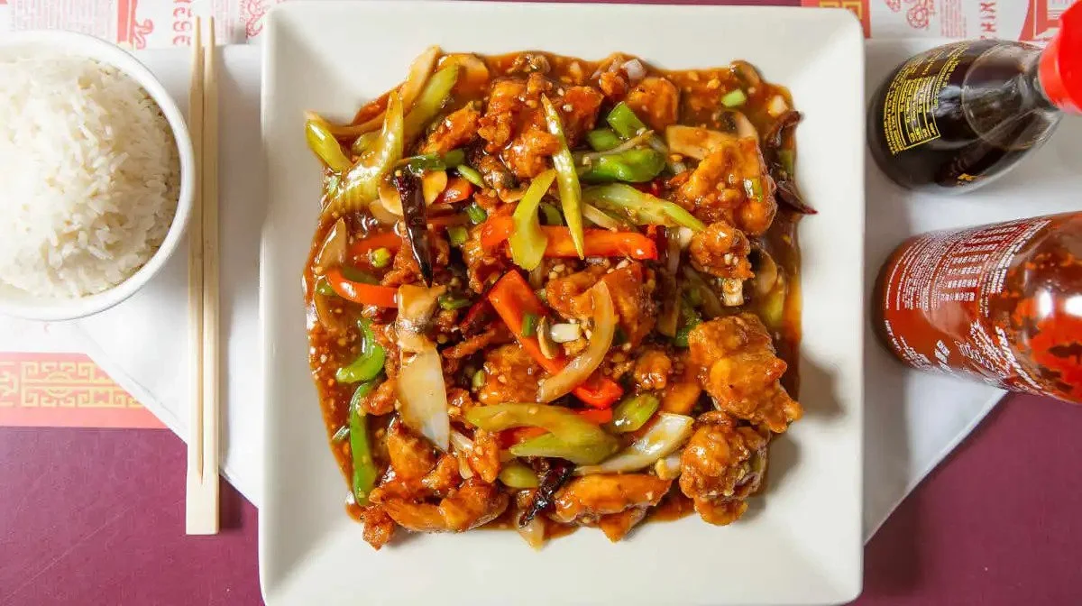 Plate of orange chicken with mixed vegetables, bowl of white rice, and soy sauce and hot sauce bottles on a table with chopsticks.