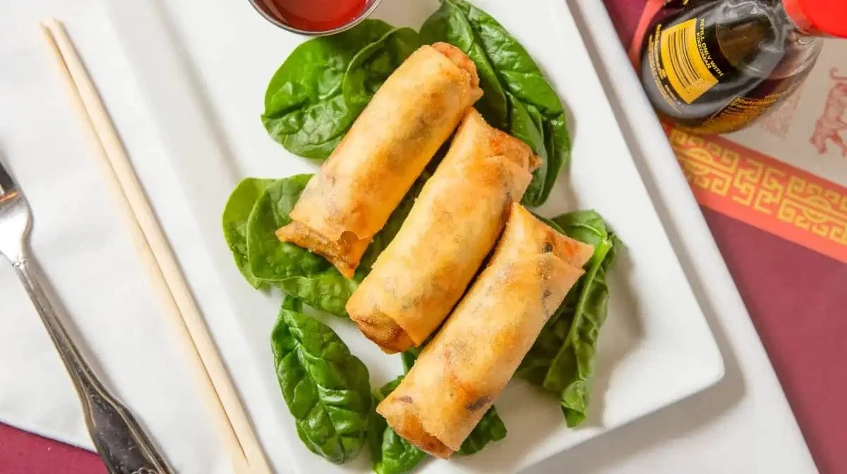 Three spring rolls on a white rectangular plate garnished with fresh spinach leaves, with soy sauce and a small bottle of soy sauce in the background.