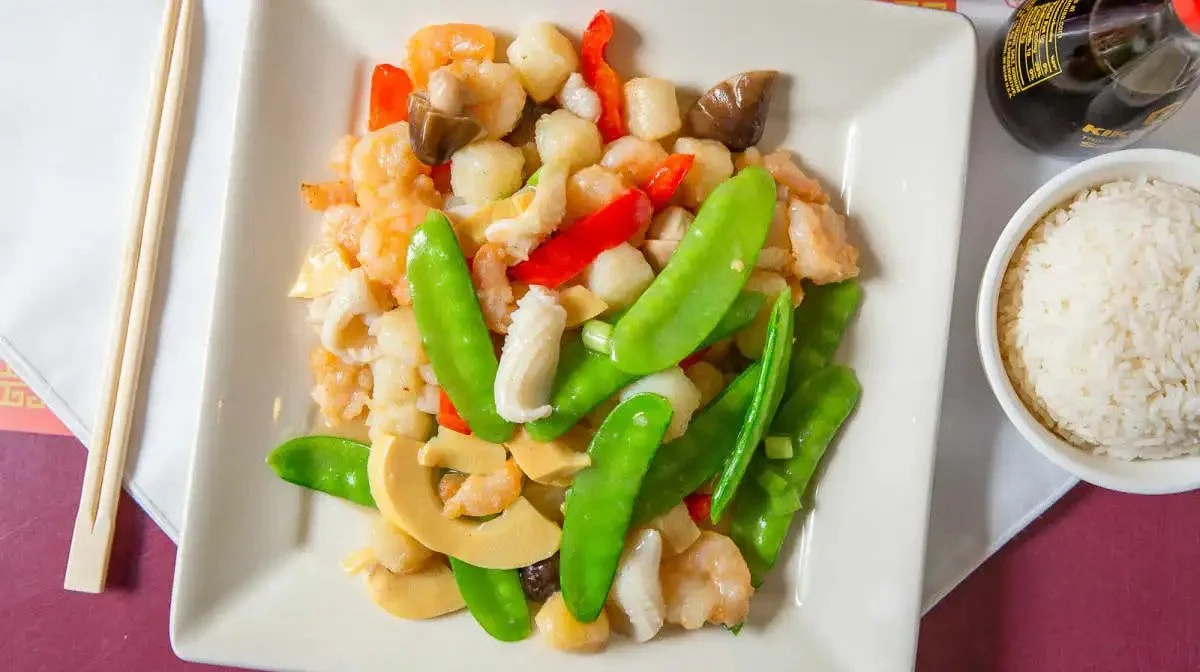 Stir-fried mixed vegetables and seafood served on a white square plate with a bowl of white rice and a bottle of soy sauce on the side