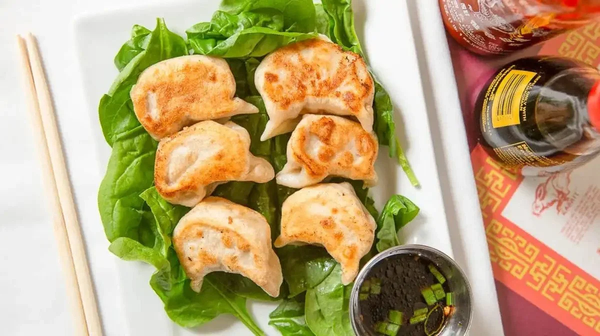 Six fried dumplings on a white rectangular plate with green lettuce in the background. Accompanying the dumplings is a small dipping bowl of soy sauce with chopped green onions. To the right are two bottles of soy sauce with red and yellow labels, pa