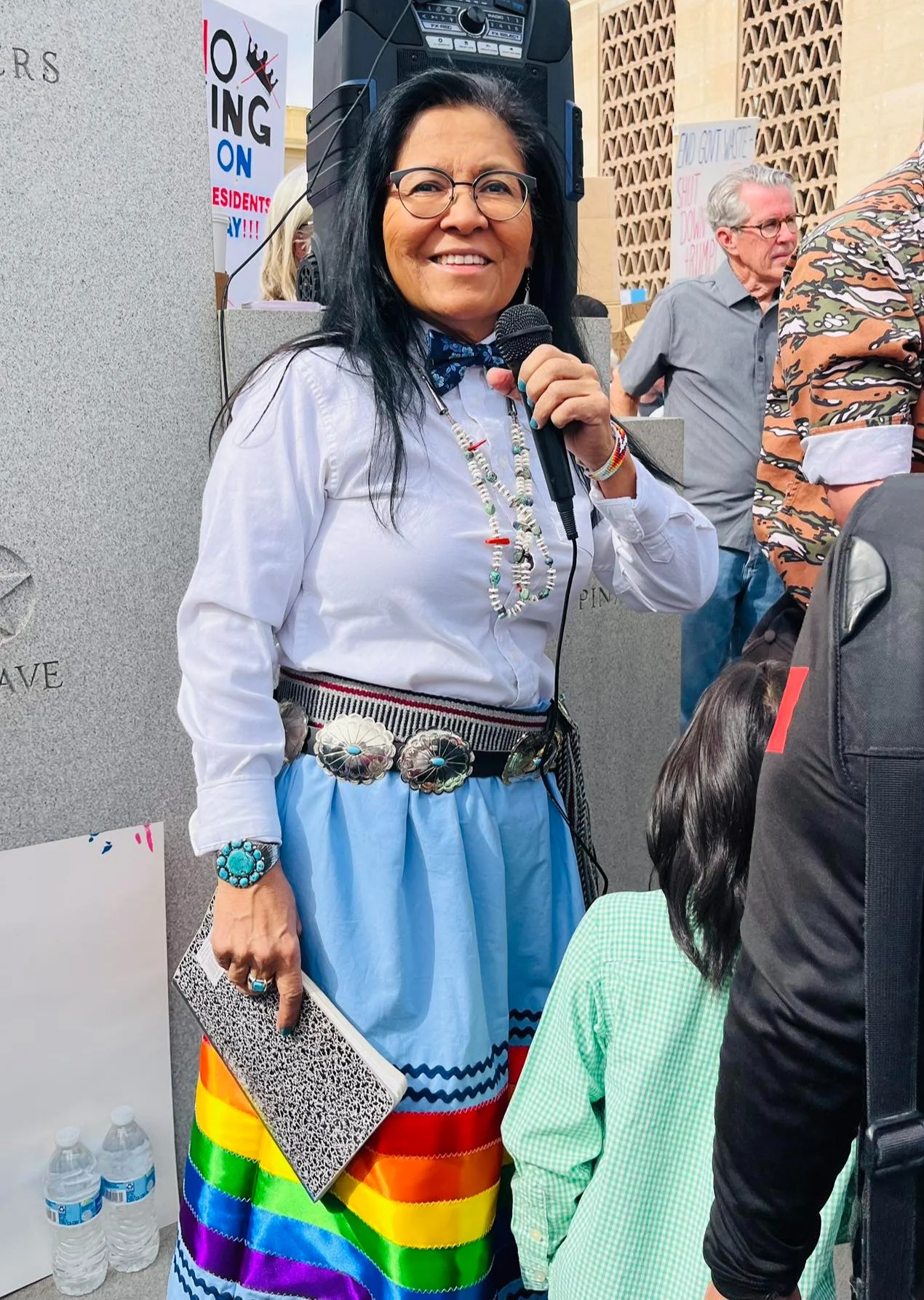 A woman with long black hair, glasses, and a white shirt smiling while holding a microphone at a protest or rally. She is wearing a blue skirt with rainbow-colored stripes and synthetic jewelry, and is surrounded by people, some of whom are partially visible. There are signs and a speaker in the background, indicating an outdoor event.