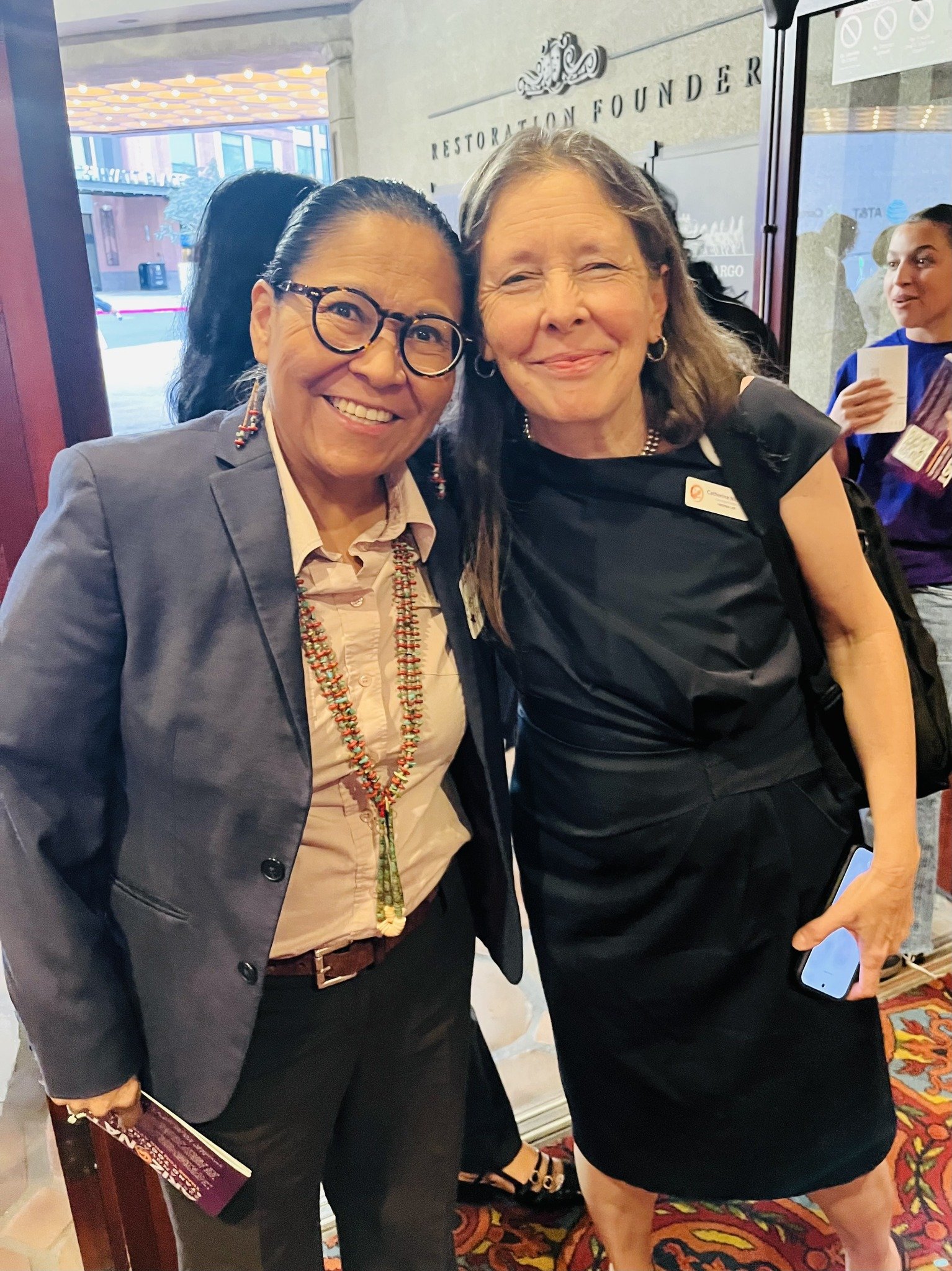 Two women smiling and posing together indoors, with a restaurant founders sign visible behind them, one with glasses and a beaded necklace, the other with a nametag and earrings.