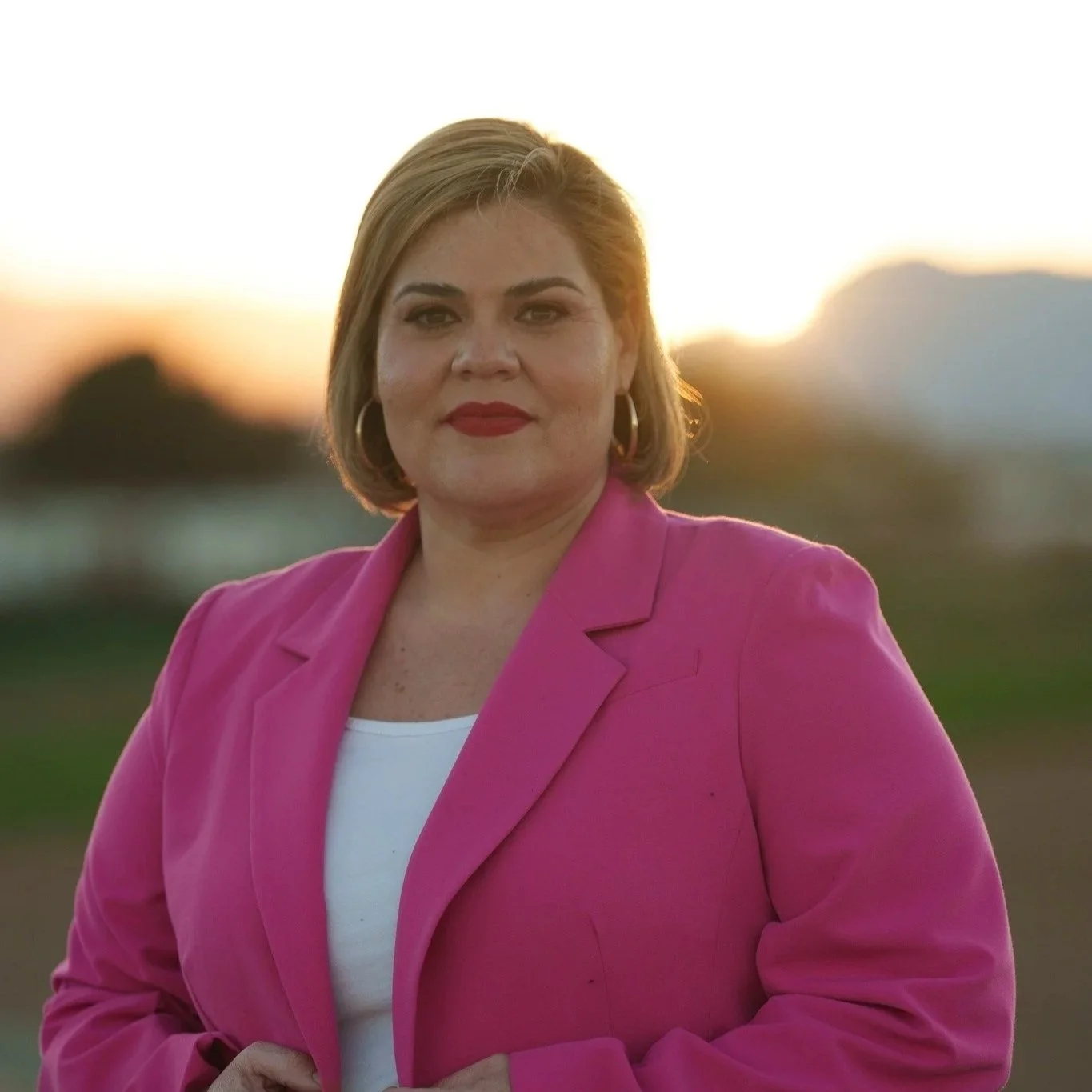 A woman with short blonde hair wearing a pink blazer and hoop earrings stands outdoors during sunset, with mountains and a blurred landscape in the background.