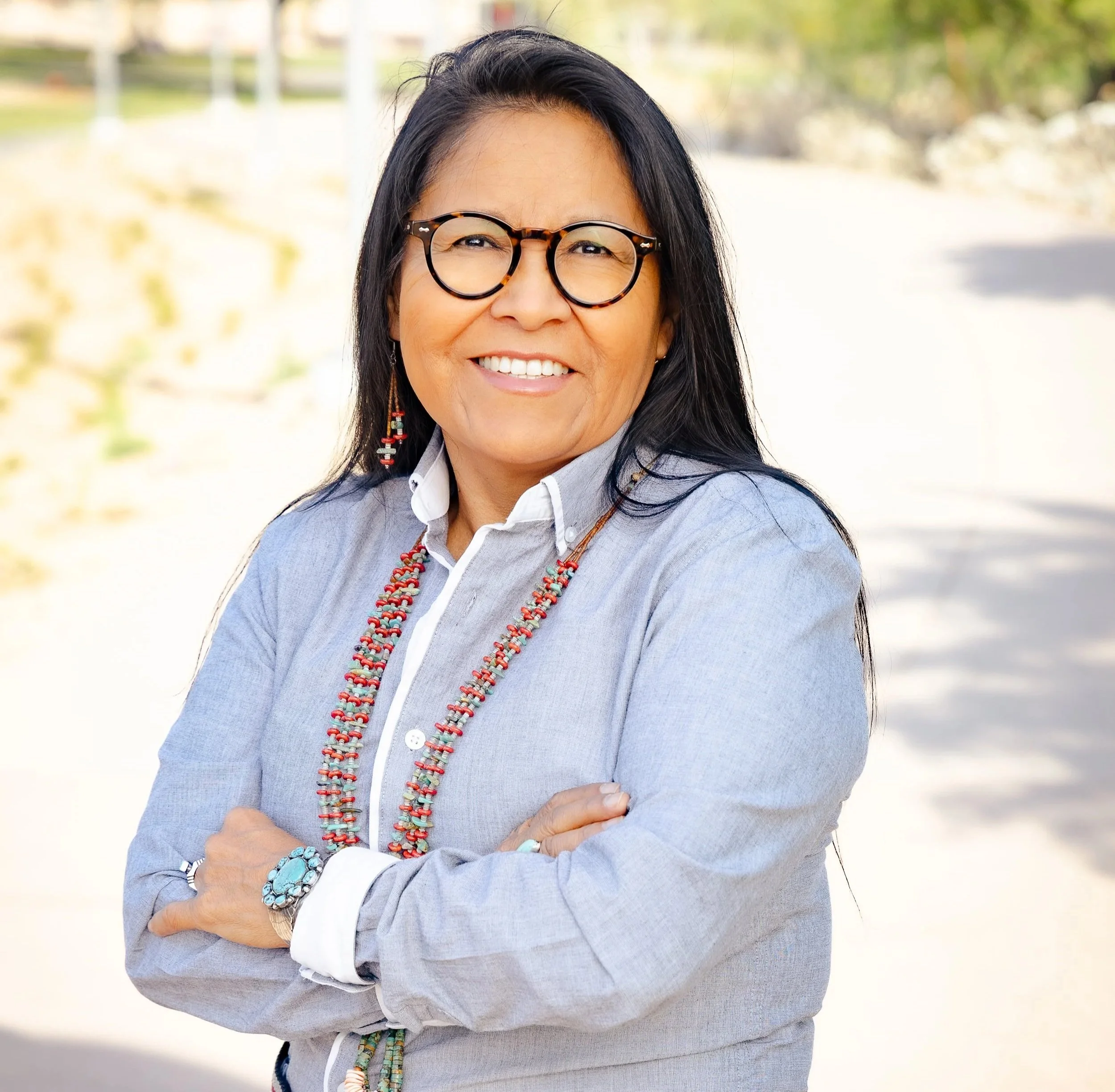 Close-up image of a woman with long black hair, wearing glasses with tortoise shell frames, smiling, outdoors with blurred trees and sunlight in the background, dressed in a light gray blazer over a white shirt, accessorized with colorful beaded necklaces and jewelry.
