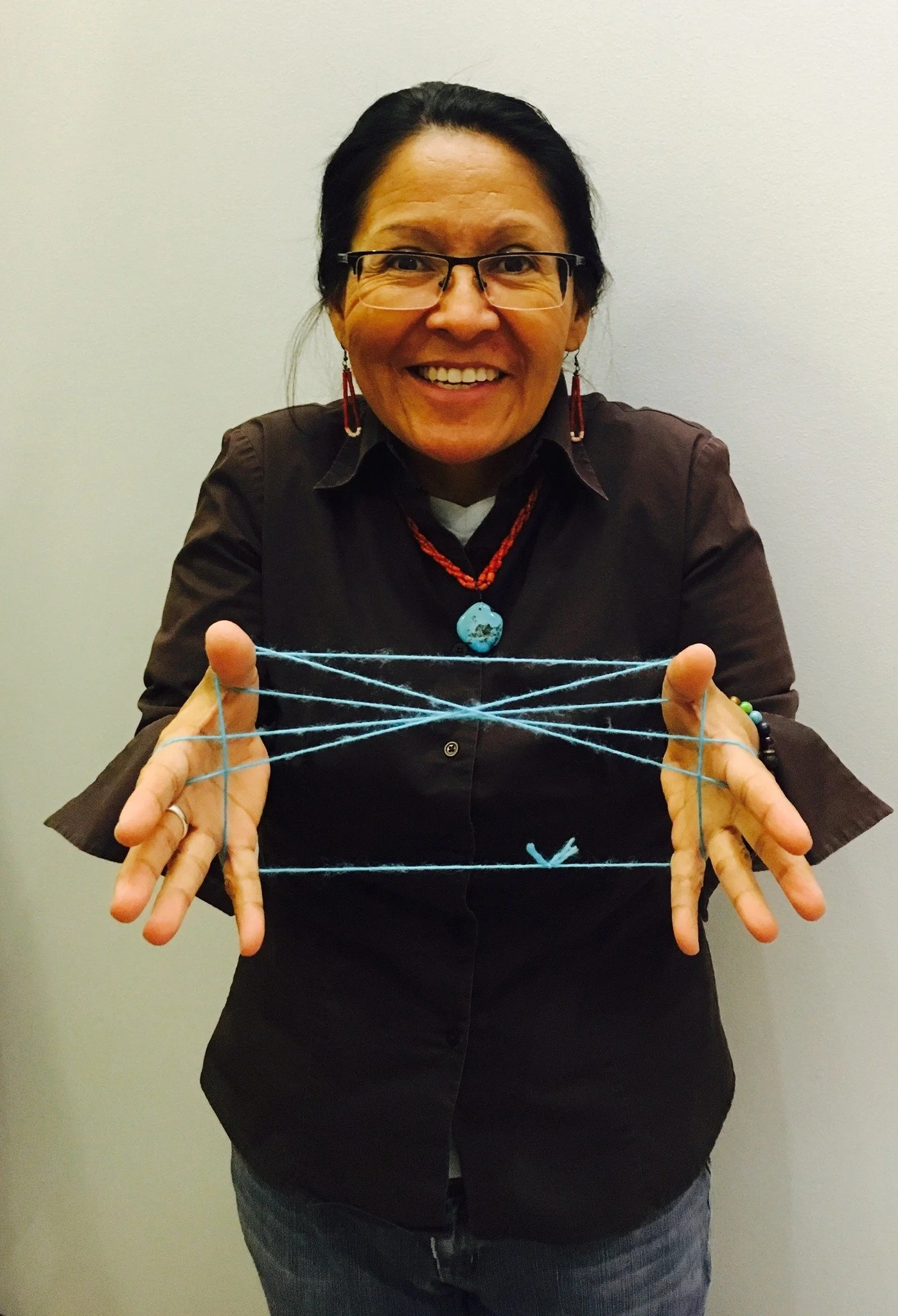 Woman smiling and holding blue yarn that is stretched across her fingers, wearing glasses, earrings, and a necklace.