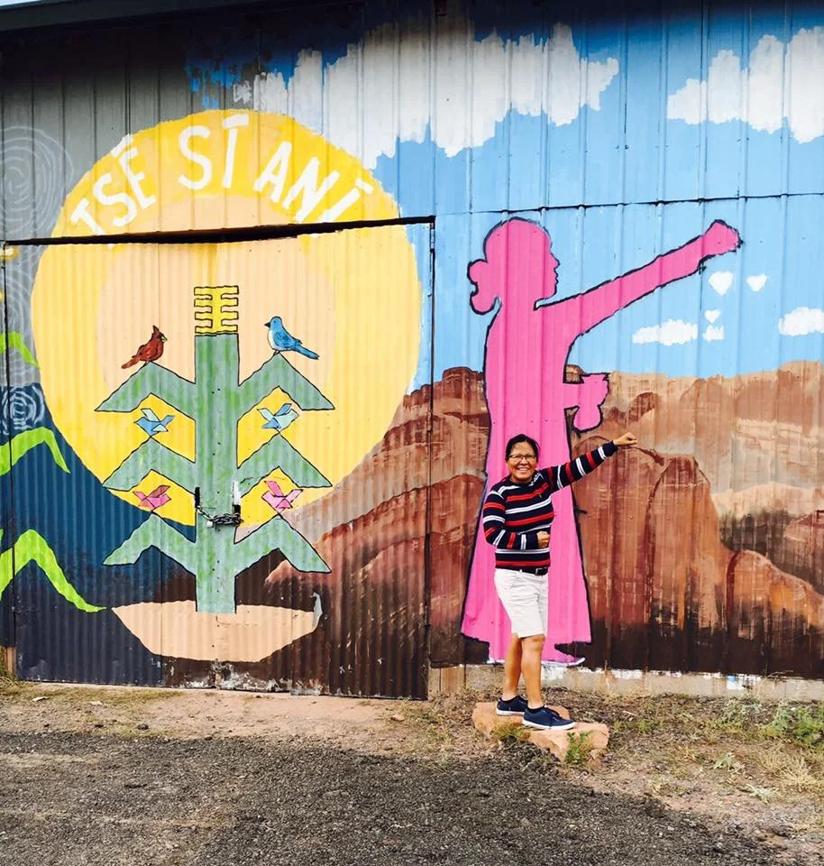 A woman posing in front of a colorful mural depicting a silhouette of a girl with outstretched arm, a mountain landscape, a tree with birds, and a yellow sun with the words "TSE STAN" partially visible.