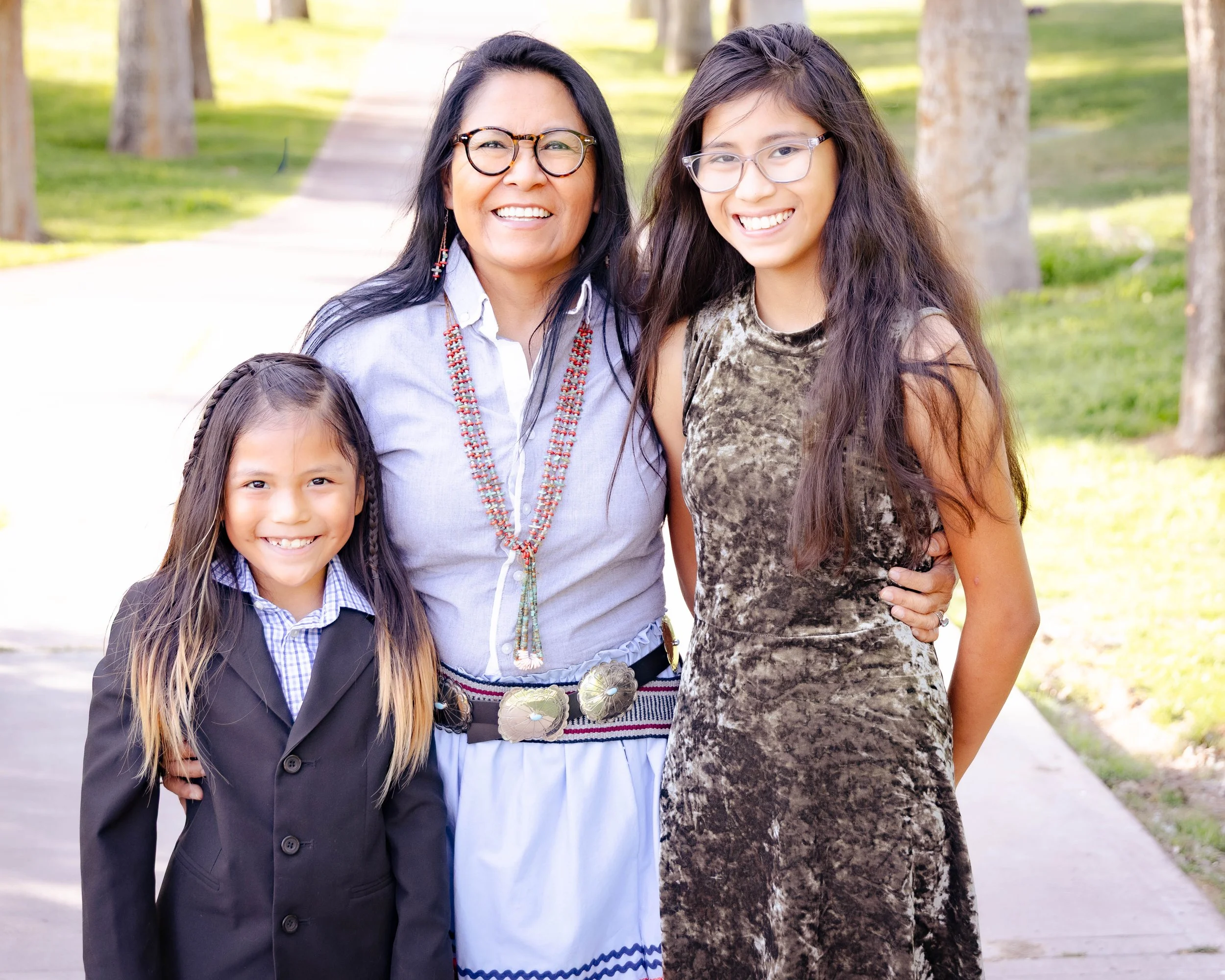 Four smiling females standing outdoors among trees, including a woman wearing glasses, a woman with curly hair, and two young girls, one in a suit and the other wearing glasses.