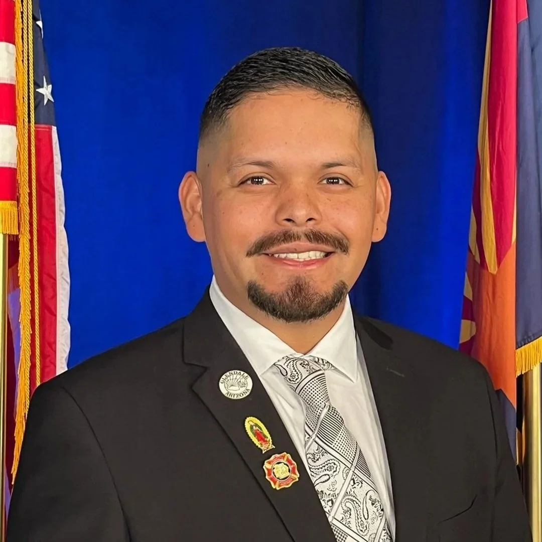 A man in a black suit with badges, white shirt, and patterned tie, standing in front of a blue backdrop with the American flag and another red, yellow, and blue flag.