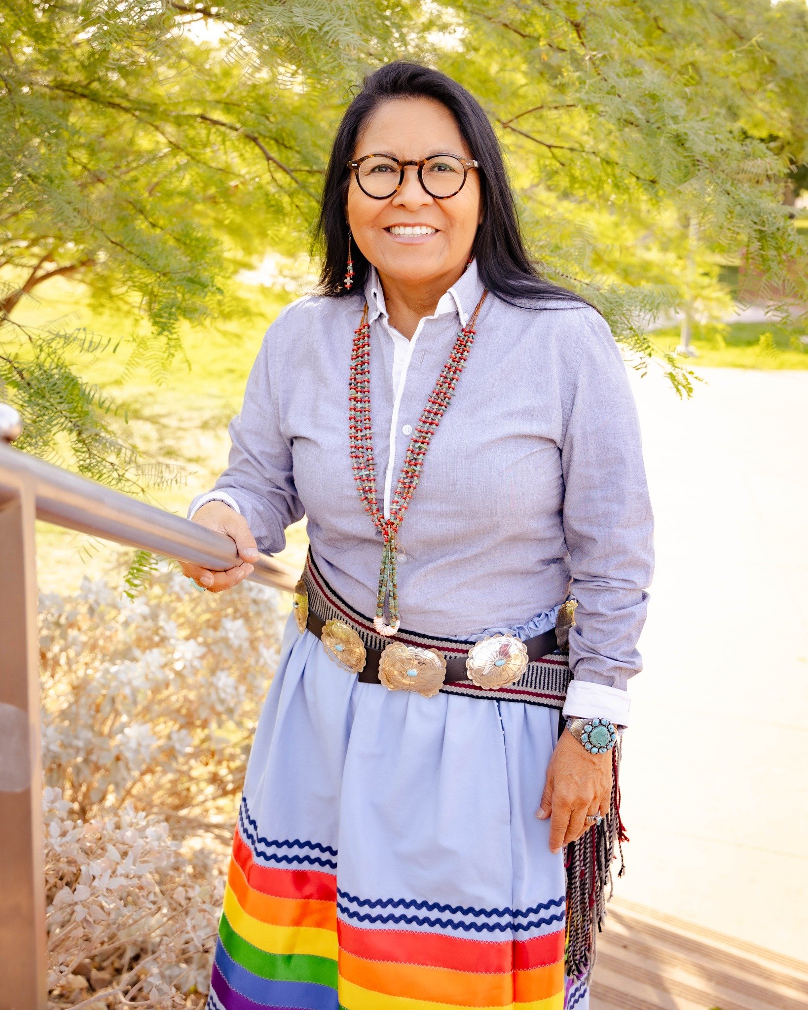 A smiling woman with black hair and glasses stands outdoors with trees and sunlight in the background. She wears a light gray shirt, colorful jewelry, and a skirt with rainbow and national flag colors, holding onto a metal railing.