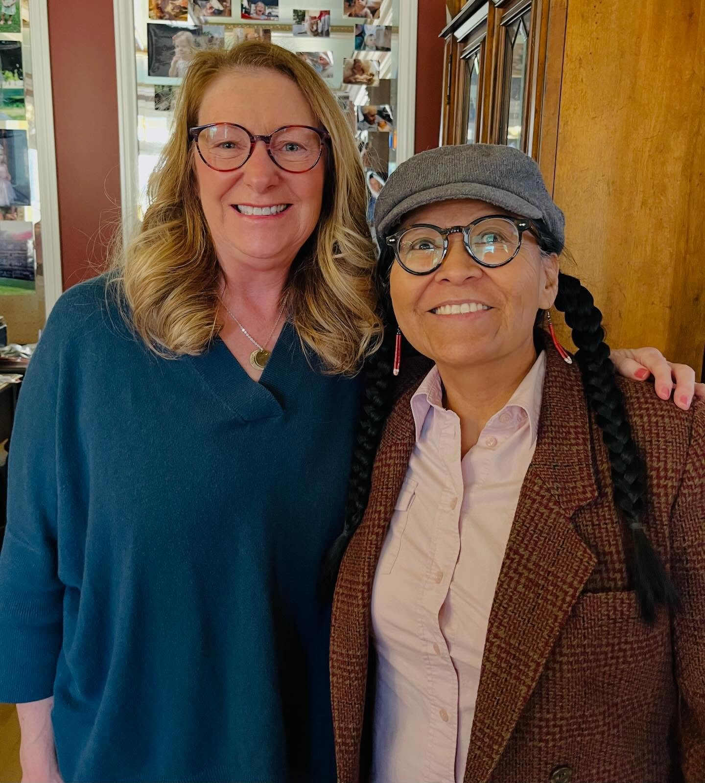 Two women smiling and posing together indoors, one with long blonde hair and glasses wearing a teal sweater, and the other with dark hair in braids wearing a gray hat, glasses, a pink shirt, and a brown plaid jacket.