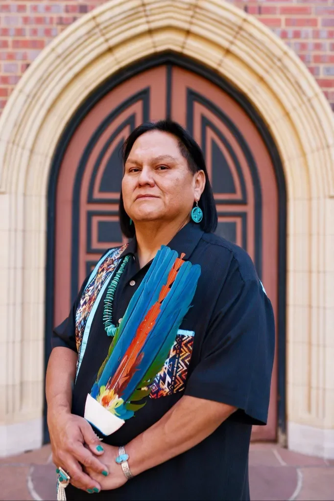 Native American woman standing in front of a church entrance, holding colorful feathers and wearing traditional jewelry and attire.