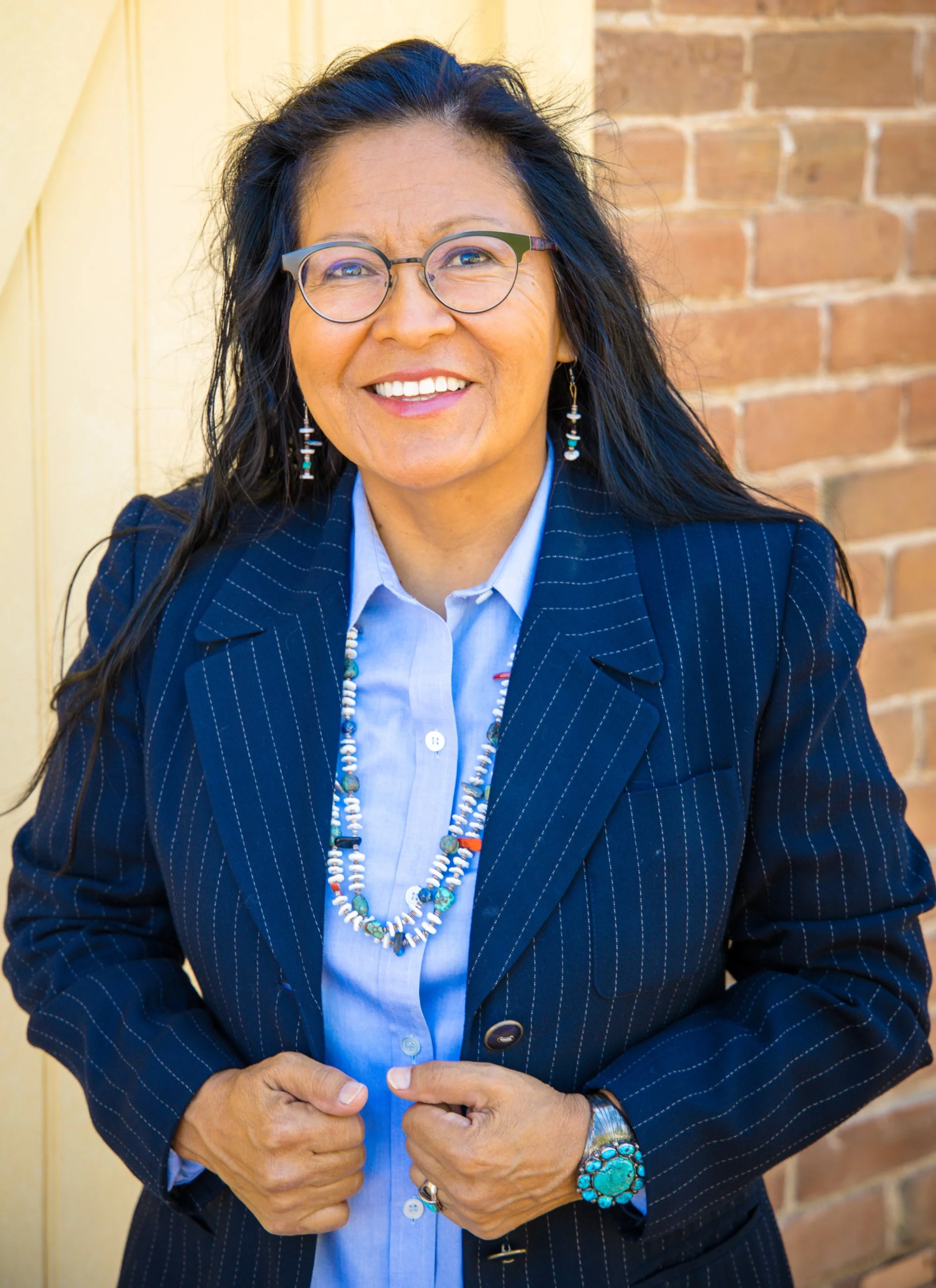 A woman with long black hair, glasses, and earrings, dressed in a blue pinstripe blazer and light blue shirt, standing outdoors near a brick wall, smiling.