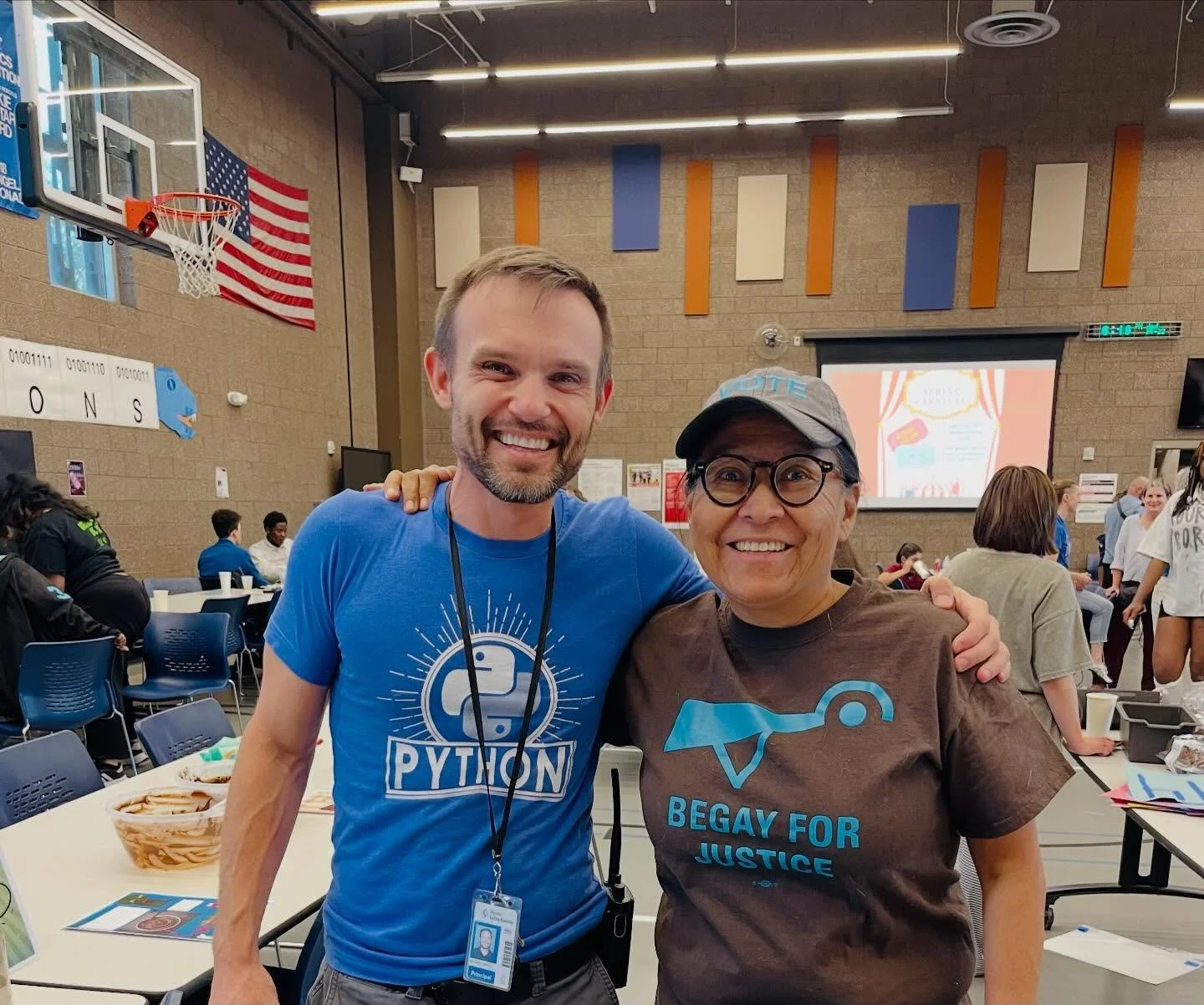 Two smiling people, one man and one woman, standing close together at a community event or gathering in a gymnasium, with tables, chairs, and other attendees in the background. The man is wearing a blue shirt with the Python programming language logo