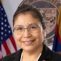 A woman with glasses, wearing a black blazer, smiling in front of an American flag and a government emblem background.