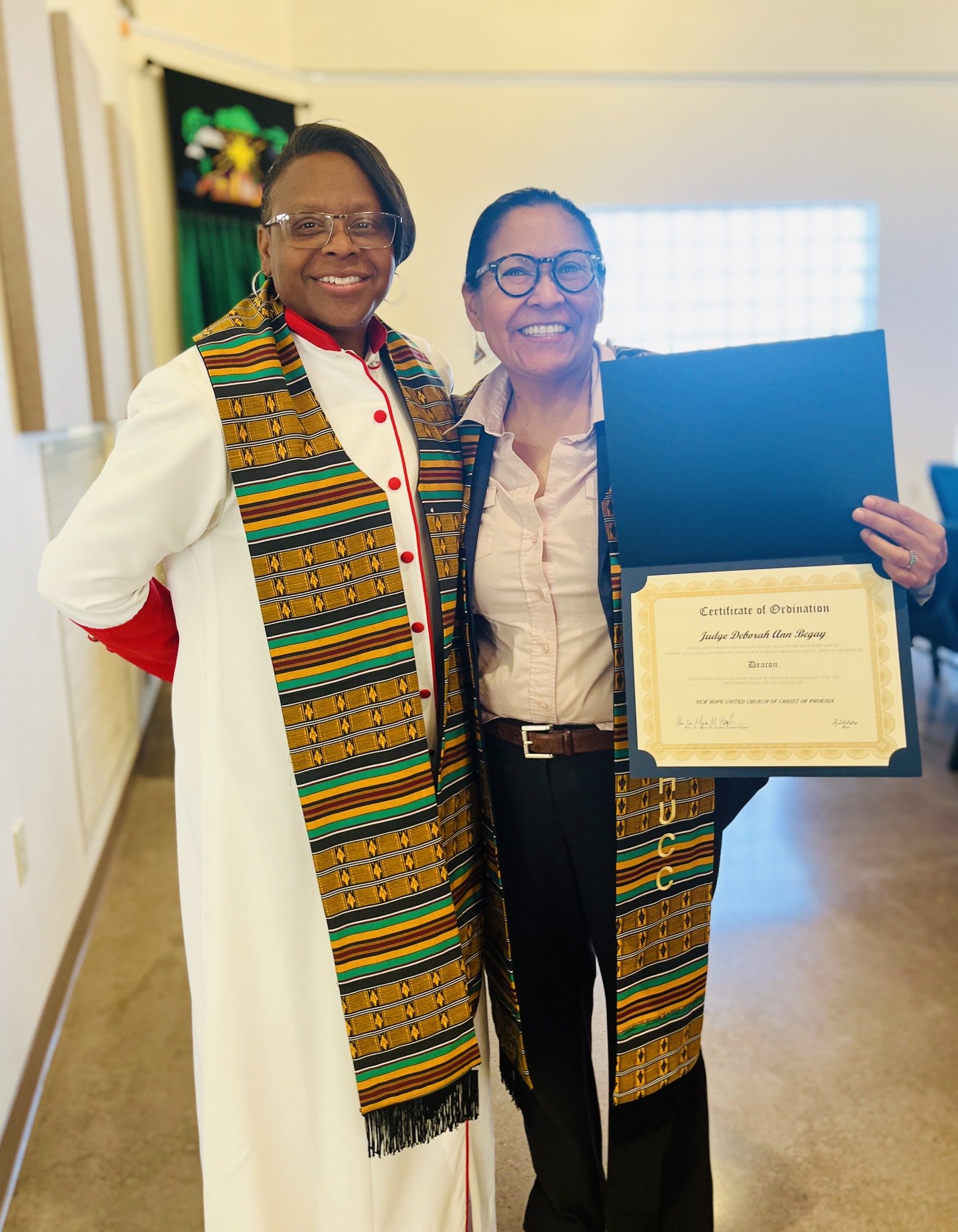 Two women standing and smiling at the camera, one holding a certificate of ordination, wearing glasses, and a patterned sash, in a room with light-colored walls and a display in the background.