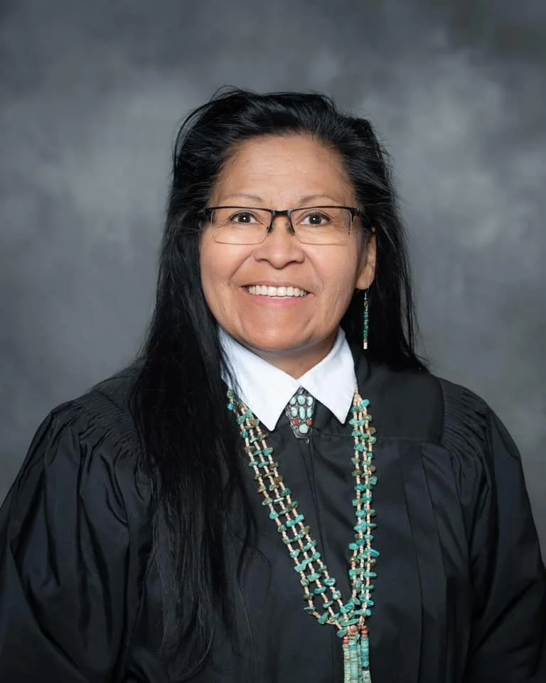 Portrait of a woman wearing glasses, a black academic gown, a white shirt, and beaded jewelry, smiling at the camera.