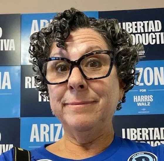 Close-up of a woman with curly gray hair and glasses, standing in front of a blue and black political campaign poster.