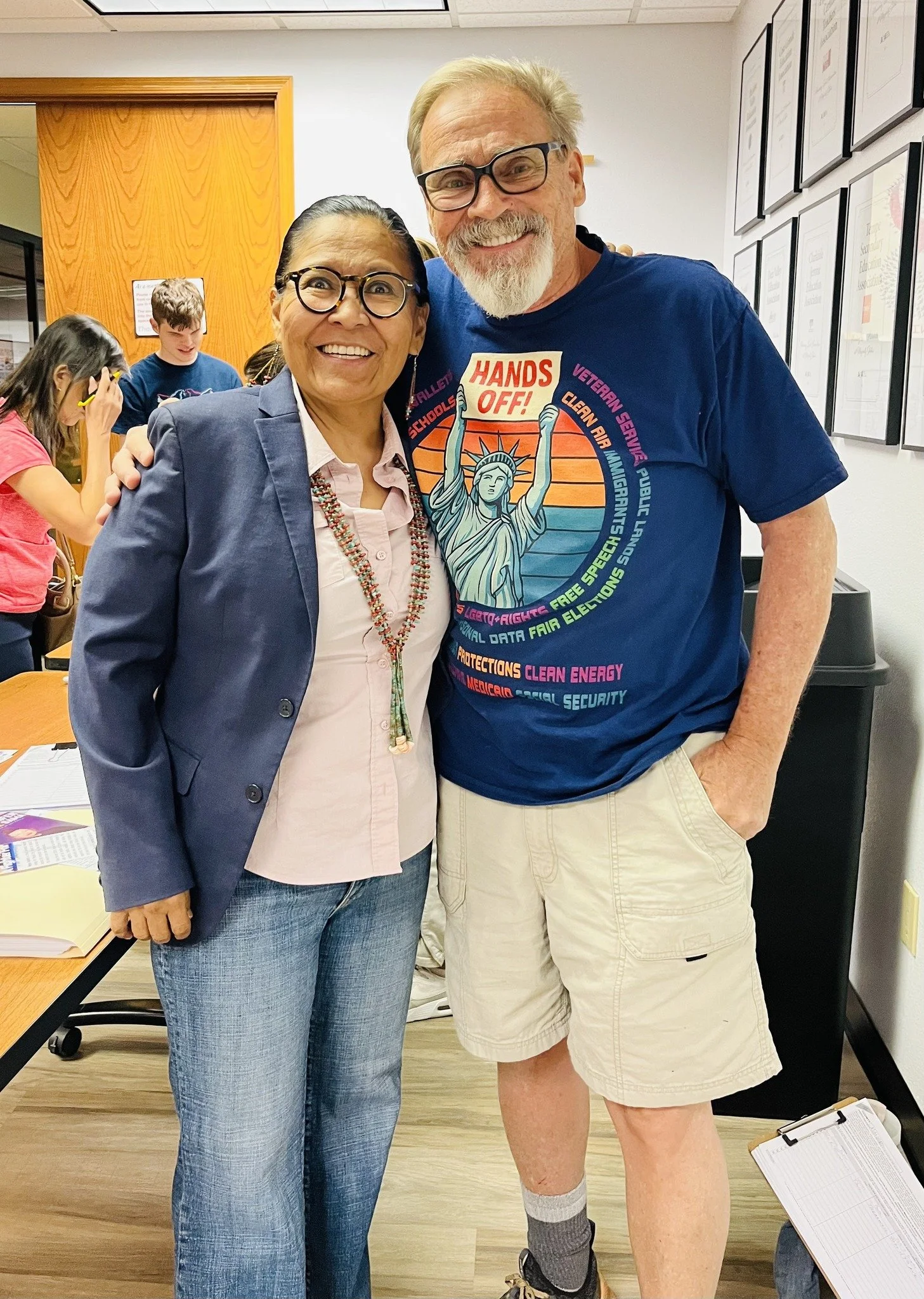 A woman and a man standing closely together, smiling for the camera in an indoor setting. The woman has long dark hair, glasses, and is wearing a blazer and jeans. The man has a beard, glasses, and is wearing a colorful t-shirt, shorts, and socks wit