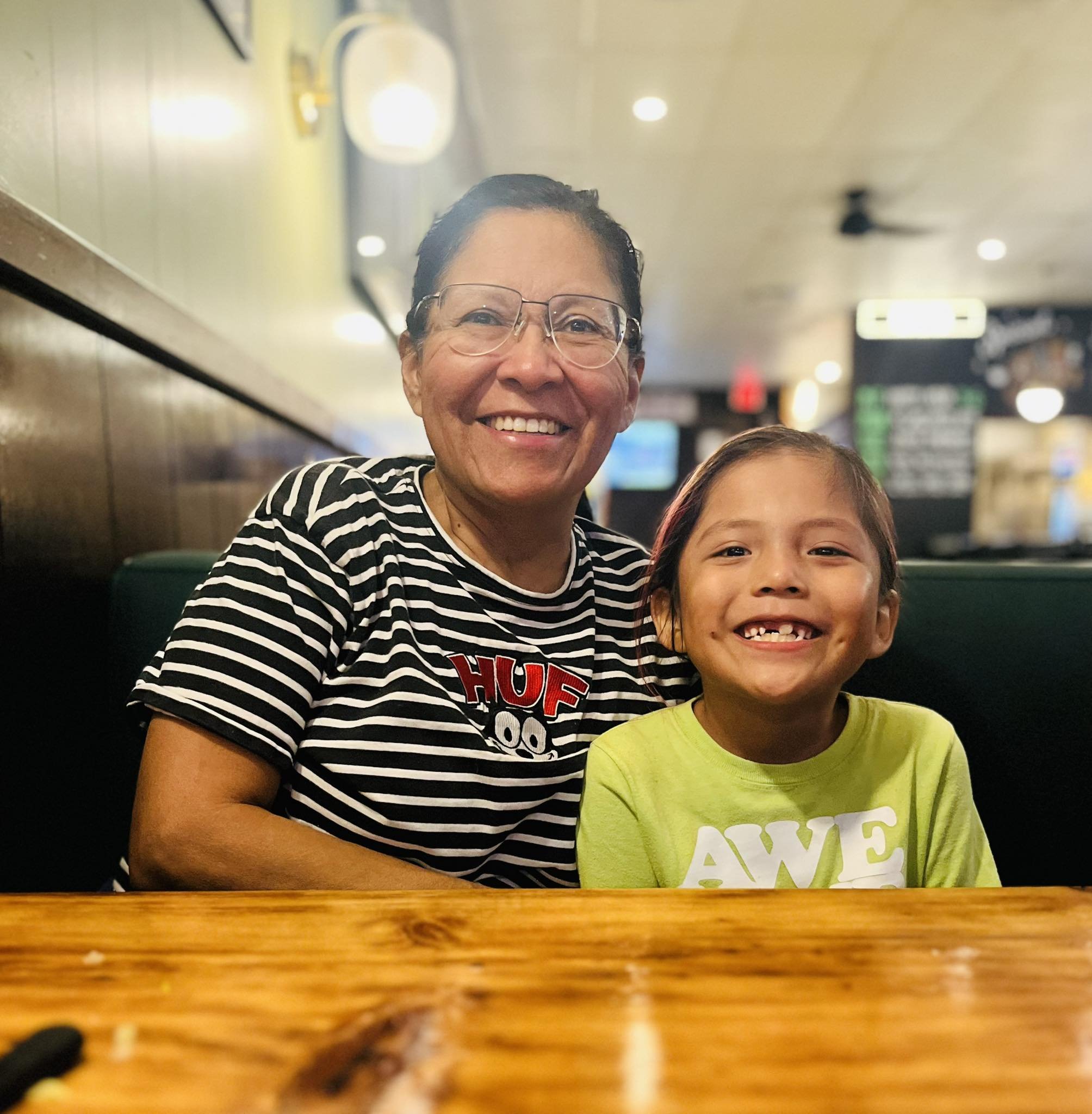 Smiling woman with short dark hair and glasses, and a young girl with brown hair and a bright yellow shirt, sitting at a wooden table in a restaurant or cafe.