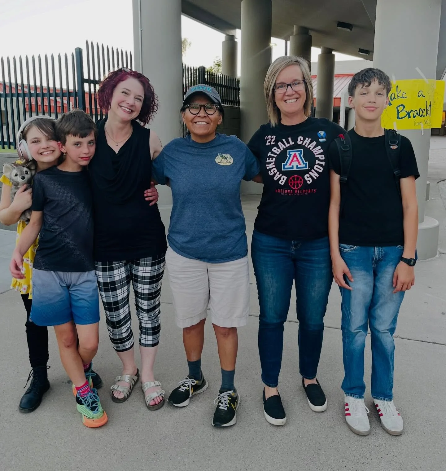 Group of six people standing outdoors, smiling for a photo. There are three women and three children, with a sign in the background that says 'Make a Bracelet.'