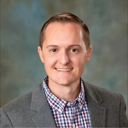 Headshot of a man with short light brown hair, wearing a gray blazer and a checkered shirt, smiling against a blurred background.