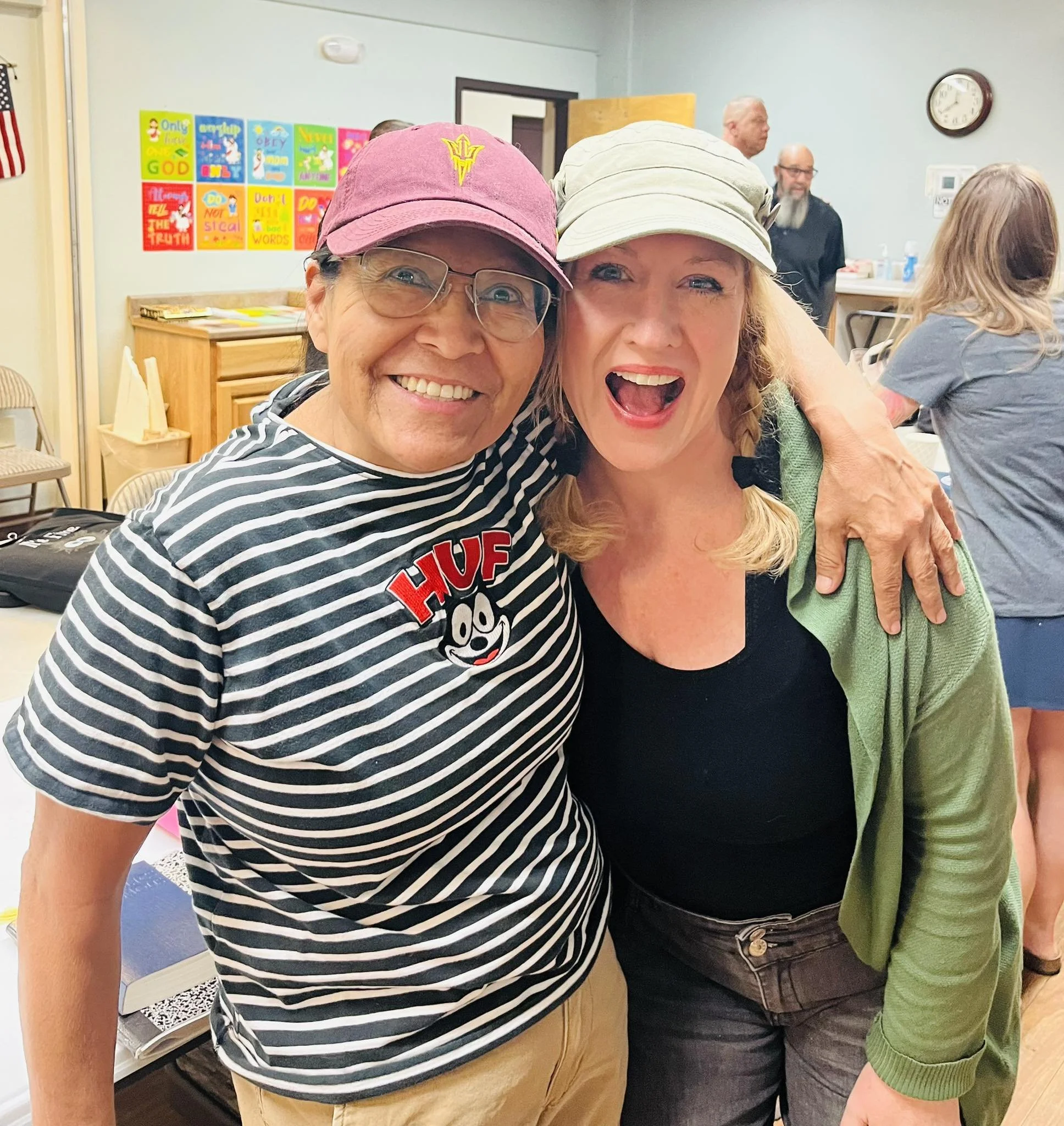 Two women are taking a selfie together at an indoor gathering. The woman on the left is wearing glasses, a pink baseball cap with a yellow emblem, and a black and white striped shirt. The woman on the right has red hair, wears a beige cap, and a blac