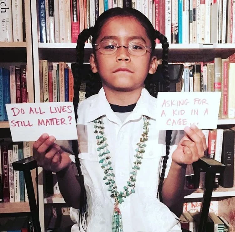 A young girl with braided hair wearing glasses and a white shirt, sitting in front of a bookshelf. She holds two signs: one that says 'Do all lives still matter?' and another that says 'Asking for a kid in a cage...'