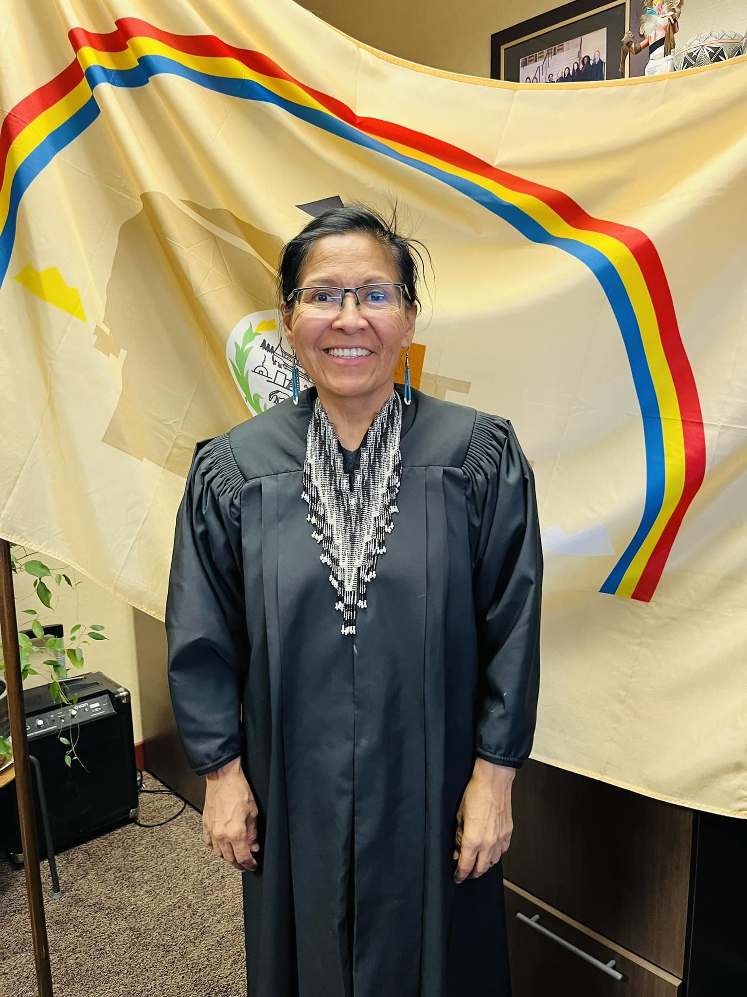 A woman wearing glasses and a black gown stands in front of a large flag with red, yellow, and blue stripes. She is smiling and has earrings and a beaded necklace. The background includes a framed photo on the wall and some decorative items.