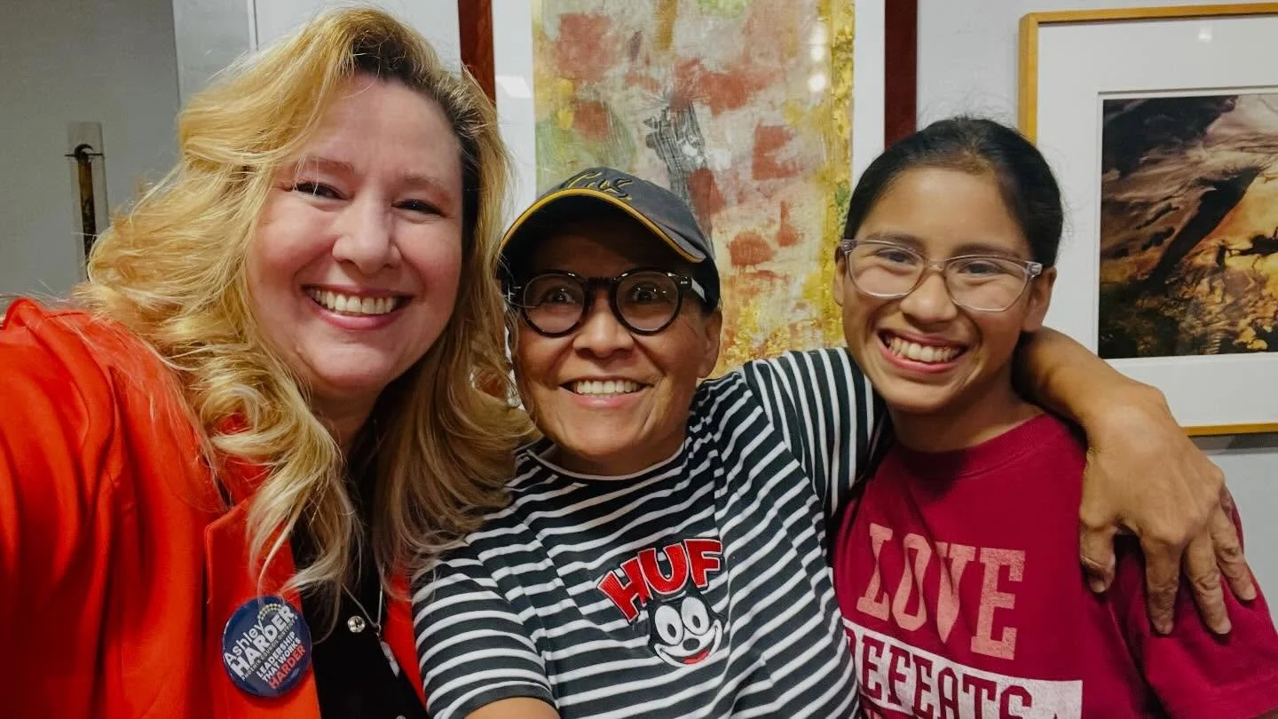 Three women smiling and hugging each other, standing indoors in front of colorful artwork. The woman on the left has blonde hair and wears an orange jacket, the woman in the middle has short hair, glasses, and wears a striped shirt with a comic desig