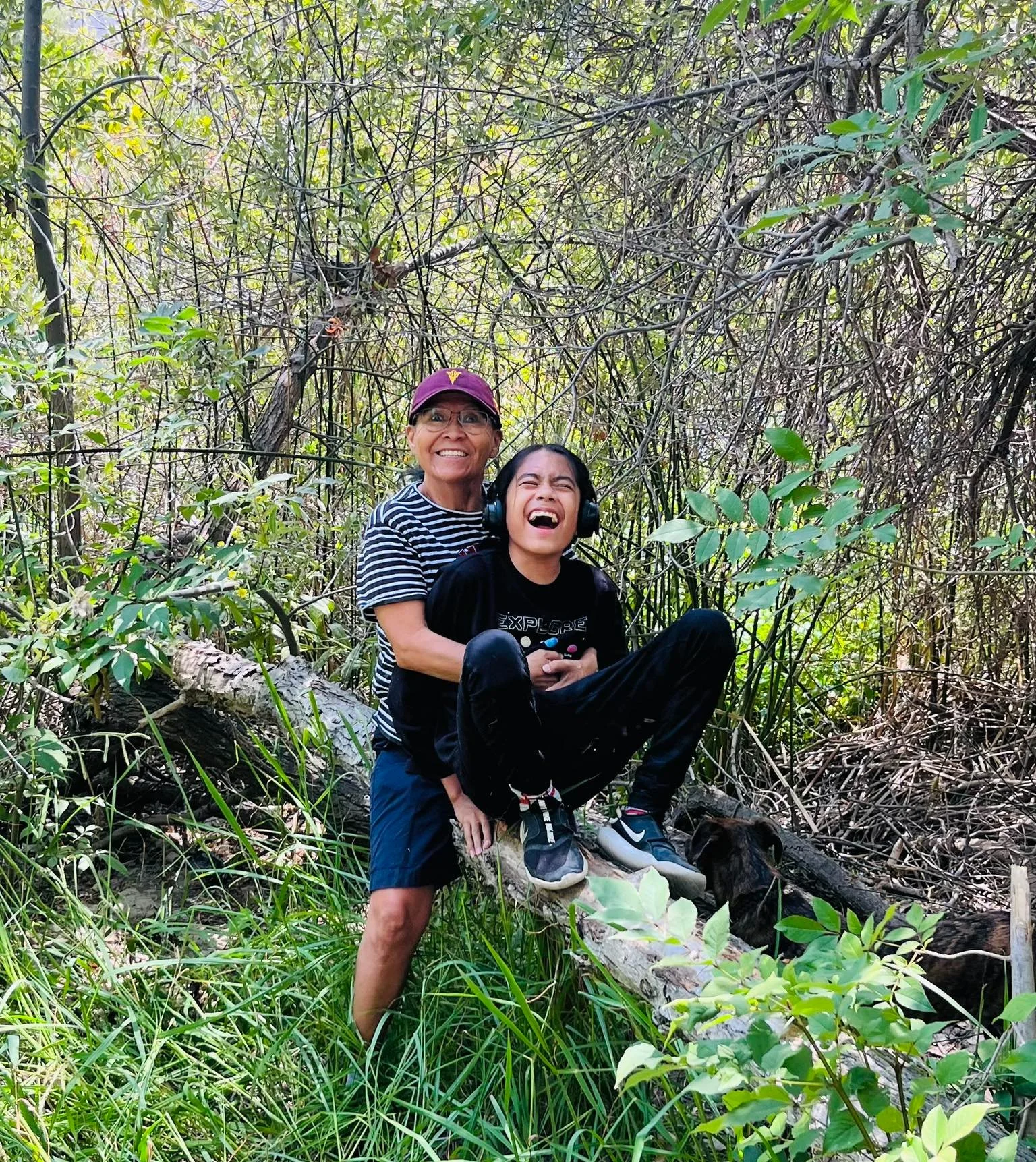 Two people, an older woman and a younger person, are in a dense, leafy forest. The woman is standing on the ground, smiling with glasses and a purple cap. The younger person, sitting on a fallen log with headphones, is laughing and leaning back into 