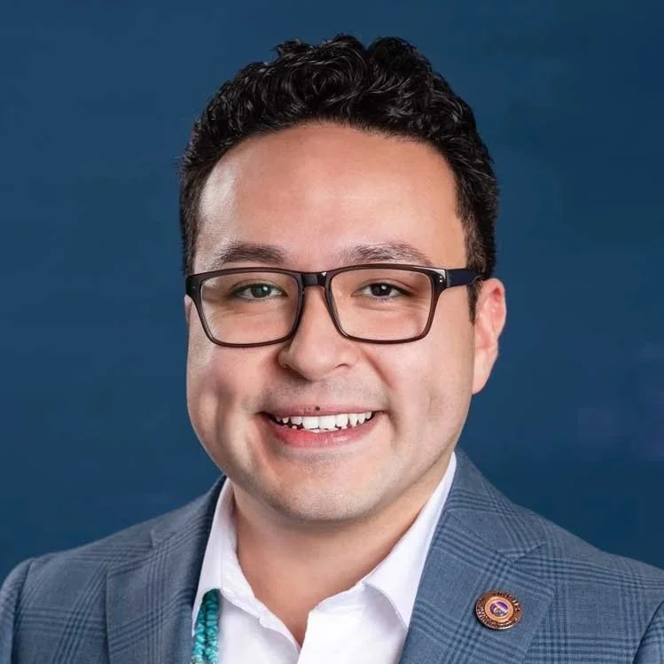 Portrait of a man with glasses, short dark hair, smiling, wearing a blazer with a lapel pin, against a blue background.