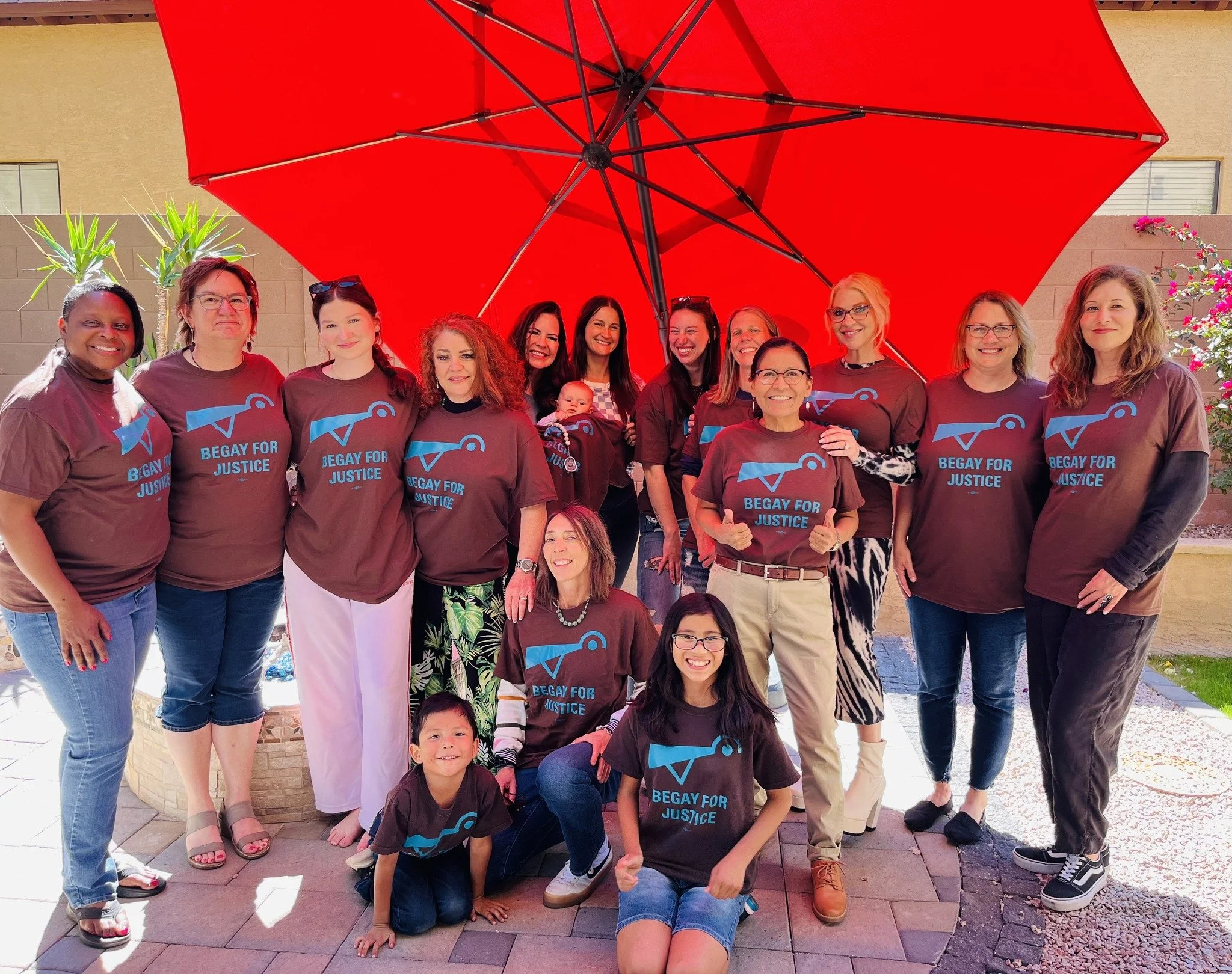 Group of people wearing matching maroon shirts with blue logos and text that says 'Begay for Justice' standing outdoors under a large red umbrella, smiling for a group photo.