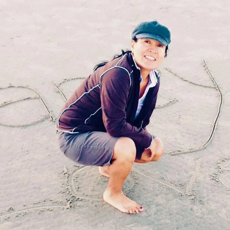 Person squatting on the beach with a smile, wearing a cap, jacket, and shorts, surrounded by palm tree sketches drawn in the sand.