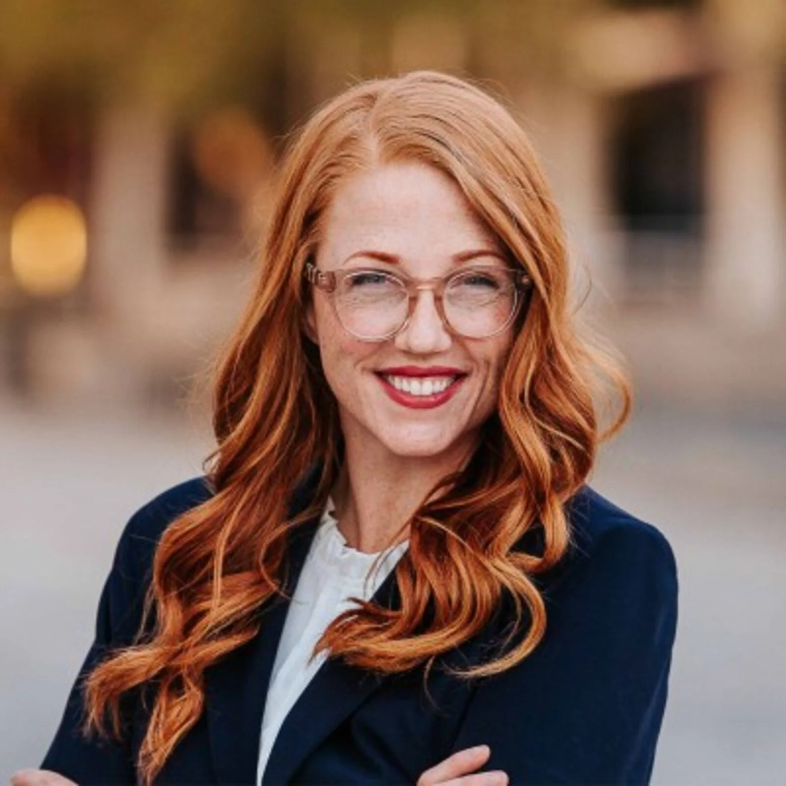 A woman with red hair, glasses, and a bright smile, standing outdoors with a blurred background.