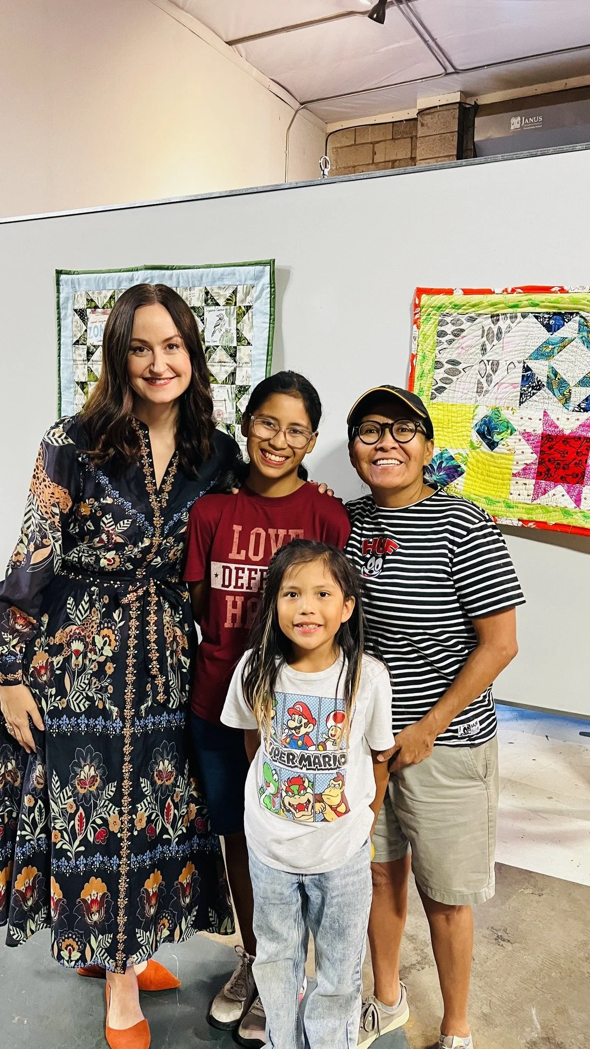Four women and a young girl stand together indoors, smiling for a photo. They are in front of a white wall decorated with colorful quilted fabric art pieces.