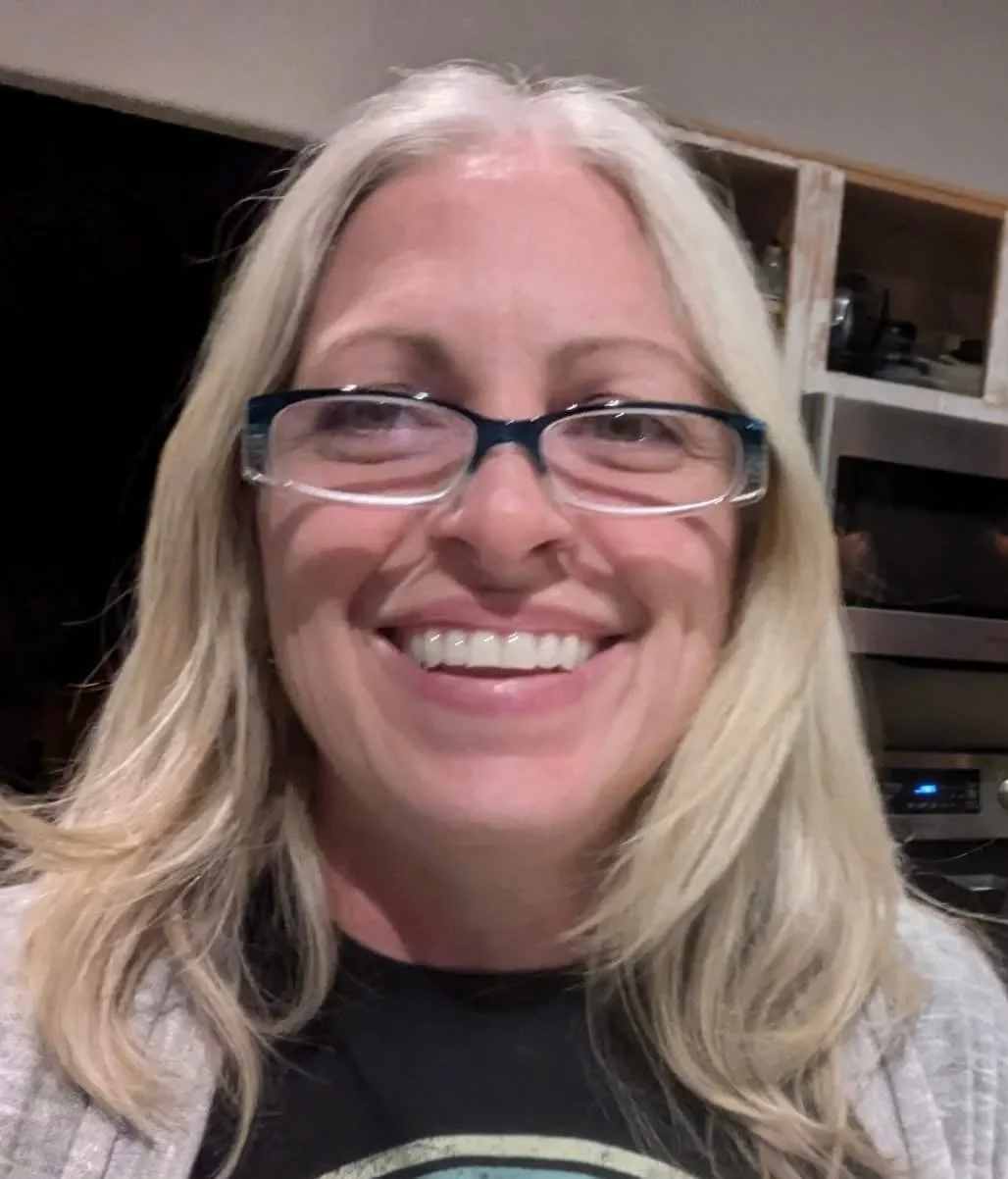 A woman with long blonde hair, wearing glasses, smiling, inside a room with kitchen shelves and appliances in the background.