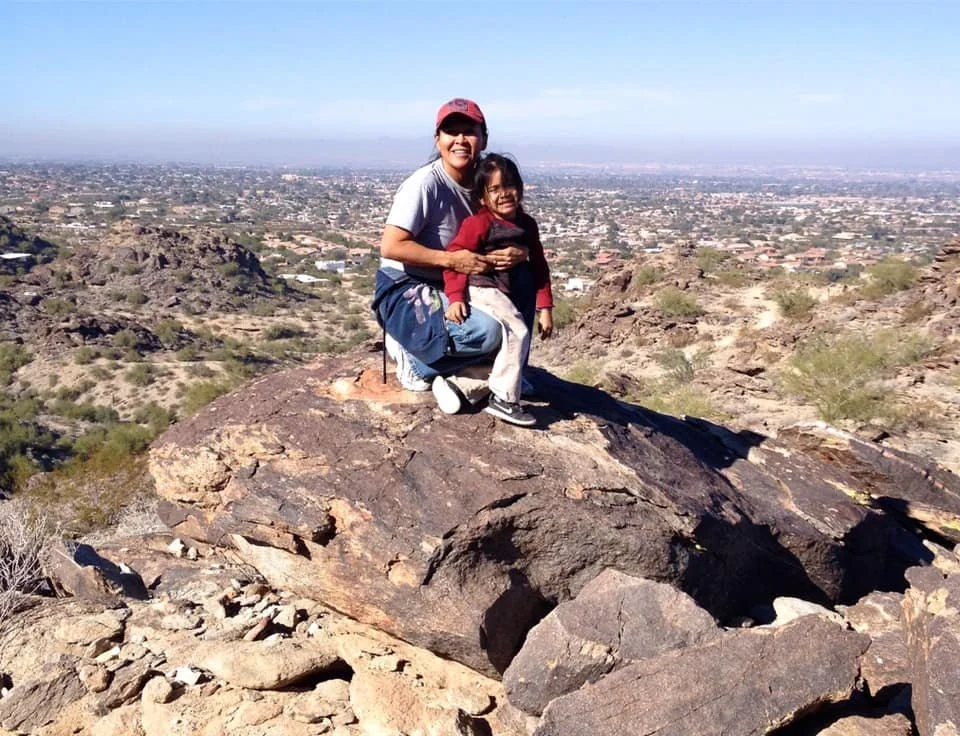 A woman and a young girl sitting on a large rock on a mountain or hilltop overlooking a cityscape, with clear blue sky overhead.