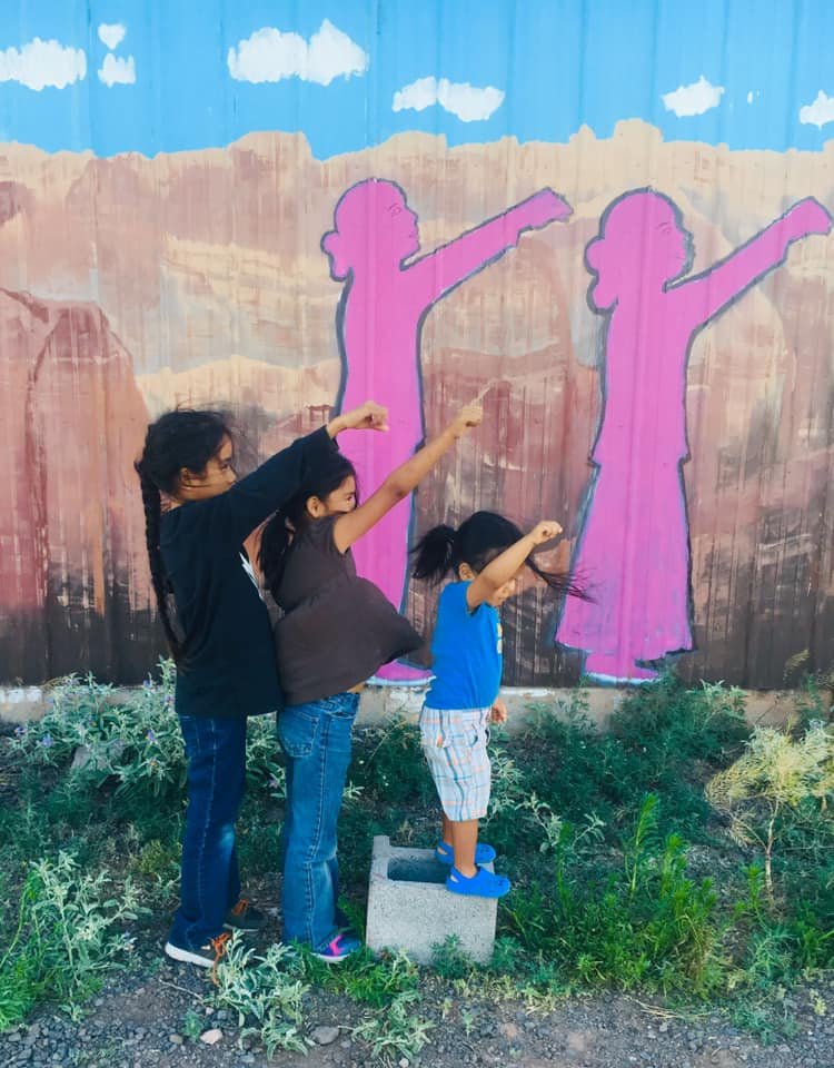 Three children standing in front of a colorful mural with pink silhouettes of girls pointing in the same direction, mimicking the poses of the figures in the mural.